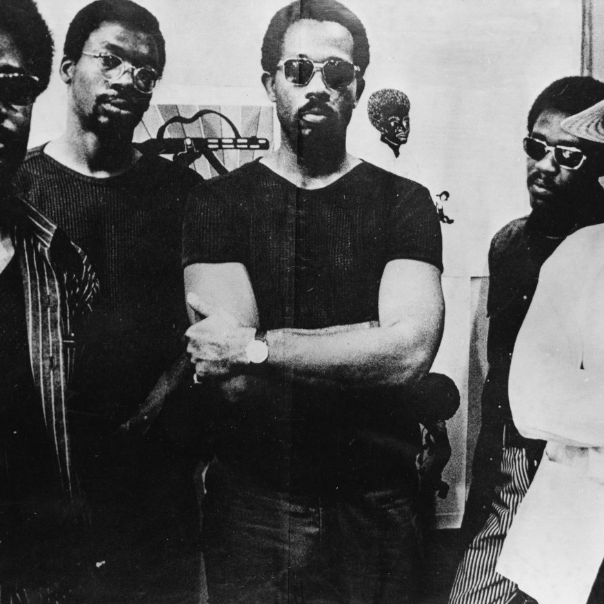 L'image montre un groupe de cinq hommes, tous portant des lunettes de soleil. Ils dégagent une certaine confiance et sont disposés côte à côte. Les hommes sont vêtus de vêtements variés, allant de t-shirts à une chemise blanche large. En arrière-plan, on peut voir des œuvres d'art abstraites et des dessins, ajoutant une dimension artistique au décor. L'ensemble de la scène dégage une atmosphère d'unité et de force, évoquant un moment important ou une rencontre significative.