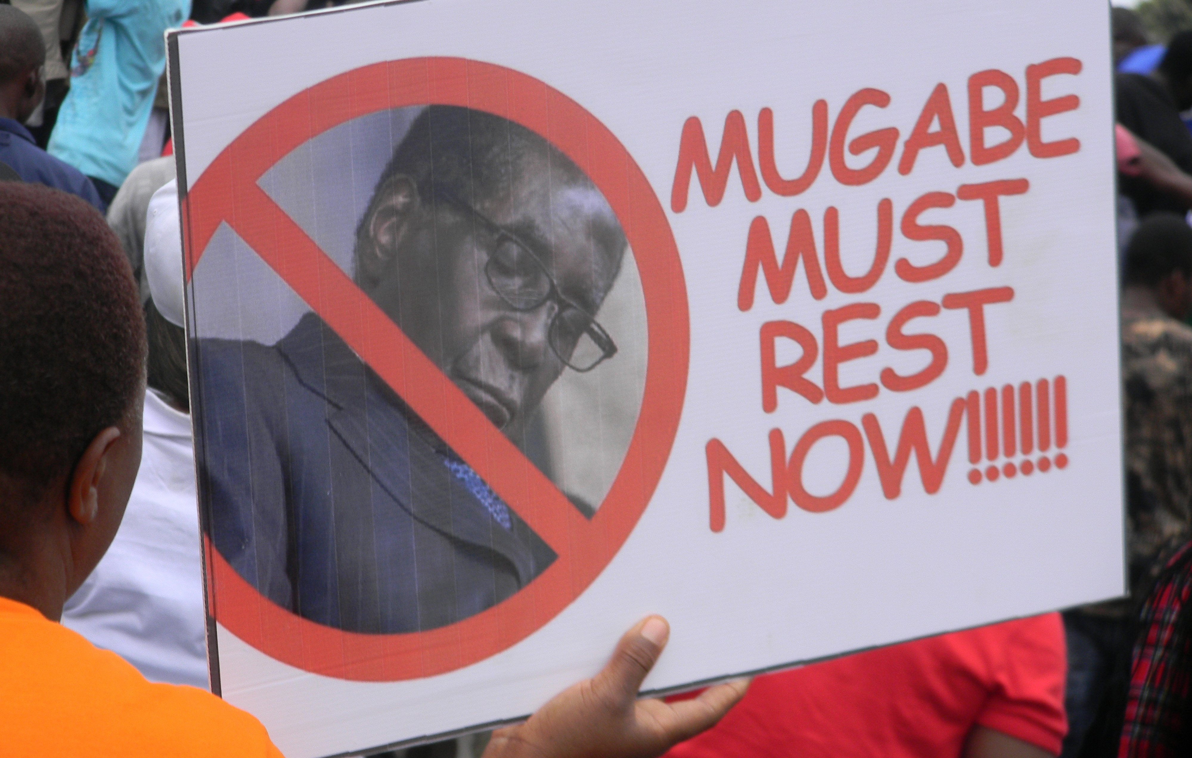 Cette image montre une personne tenant une pancarte lors d'une manifestation. La pancarte arbore un grand symbole barré, avec une photo d'un homme, généralement associé à un discours politique. Le texte sur la pancarte se lit : "MUGABE MUST REST NOW", exprimant un message clair de résistance ou de demande de cessation à l'égard de cette personne. En arrière-plan, on peut voir une foule, suggérant un rassemblement où des gens partagent des opinions fortes sur un sujet d'actualité. L'ambiance générale semble être celle d'une protestation.