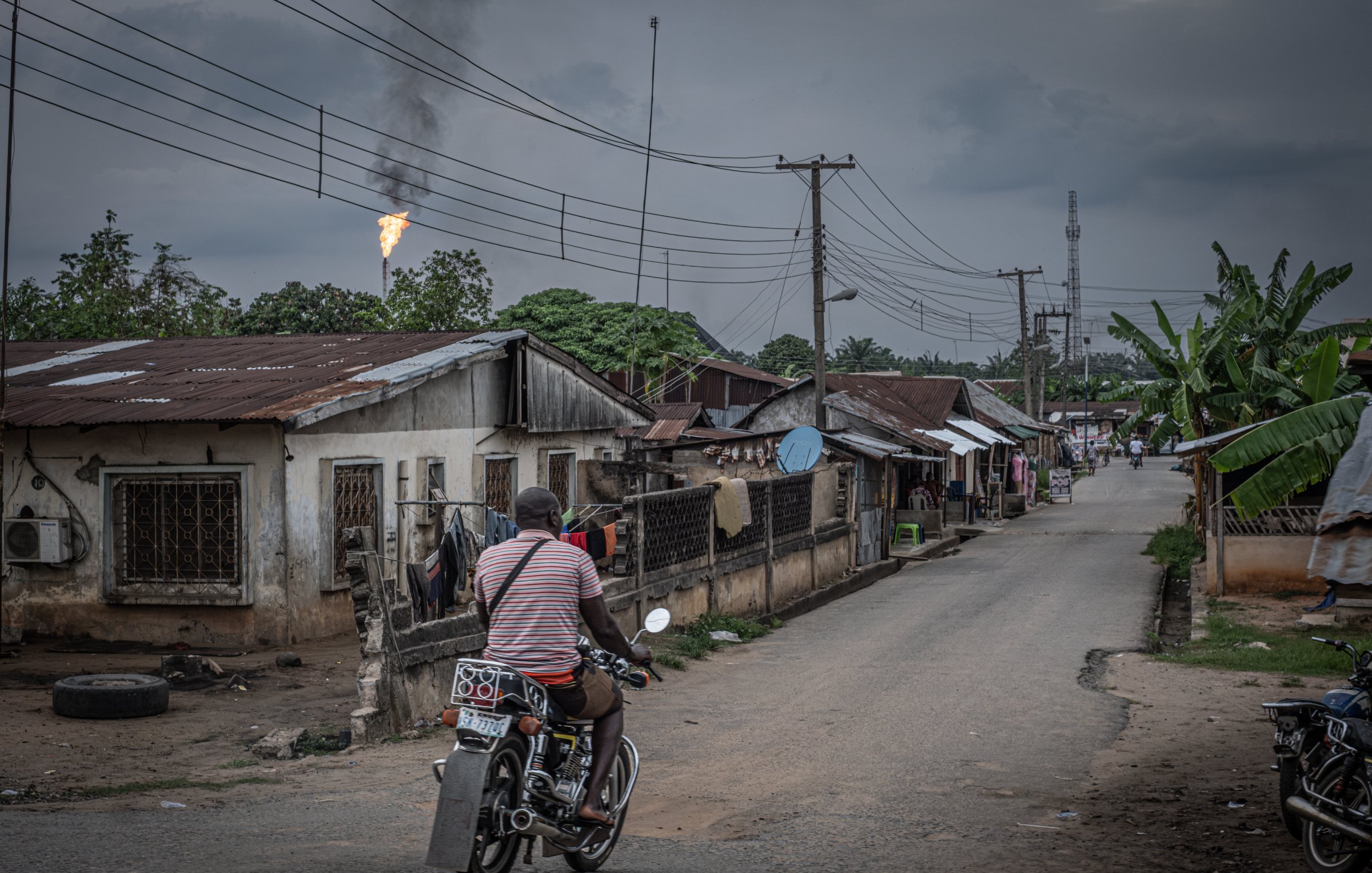 L'image présente une scène urbaine dans un quartier qui semble rural. Une route centrale est bordée de maisons modestes, souvent en tôle, avec des fenêtres grillagées. À gauche, on apperçoit un homme sur une moto, portant un t-shirt rayé, qui se dirige vers l'avant, ajoutant une touche de mouvement à la scène. En arrière-plan, des poteaux électriques sont visibles, ainsi qu'un tuyau de gaz qui rejette une flamme, indiquant peut-être une installation industrielle à proximité. Le ciel est couvert, avec des nuages sombres, créant une atmosphère légèrement mélancolique. À droite, des plantations de bananiers ajoutent une touche de verdure. L'ensemble dépeint une réalité de vie quotidienne dans un environnement à la fois simple et intéressant.