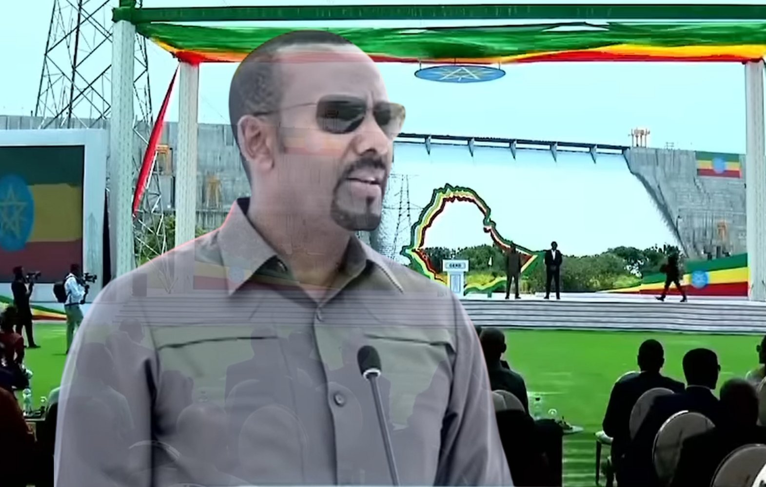 The image appears to show a man speaking at a ceremonial or public event outdoors, likely in Ethiopia. He is wearing sunglasses and has a serious expression. The background features a large stage, decorated with colorful flags, possibly representing Ethiopia. There is a crowd of people seated in front of the stage, indicating that this is a significant gathering. The event seems to be celebratory and possibly related to national pride or development.