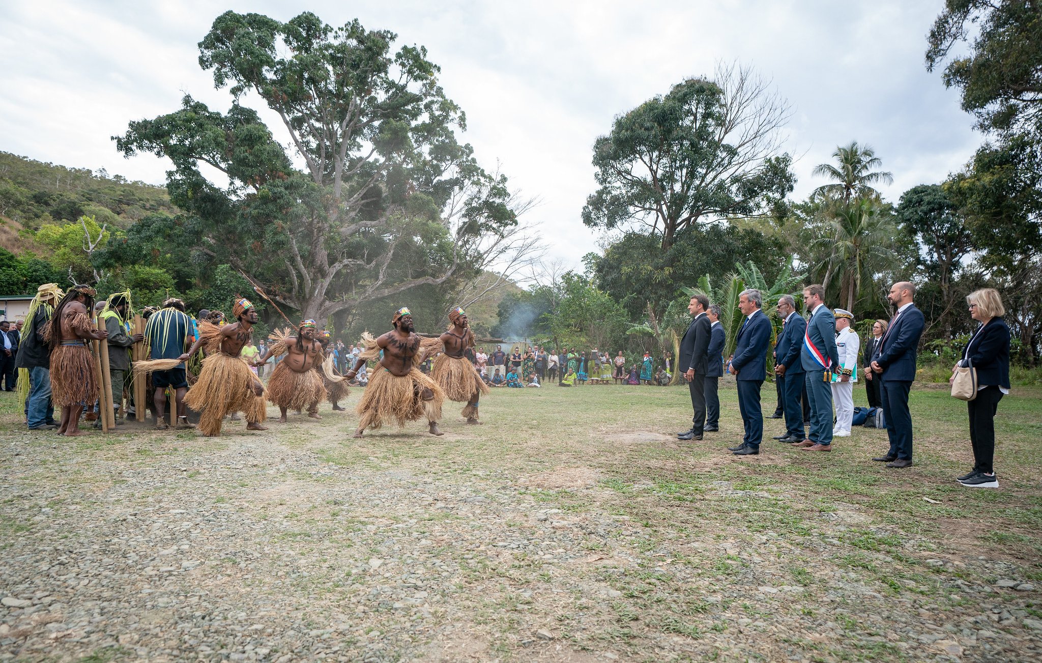 L'image montre un groupe de personnes en tenue traditionnelle, qui semblent participer à une cérémonie ou une célébration. Au centre, des hommes vêtus de costumes en fibres naturelles dansent avec énergie. Ils sont entourés par un public, composé de personnes en tenue formelle, qui observe attentivement la scène. L'environnement est verdoyant, avec des arbres et une légère brume qui donne une atmosphère presque mystique. À l'arrière-plan, un groupe de spectateurs semble apprécier l'événement, ajoutant une dimension communautaire à cette célébration culturelle.