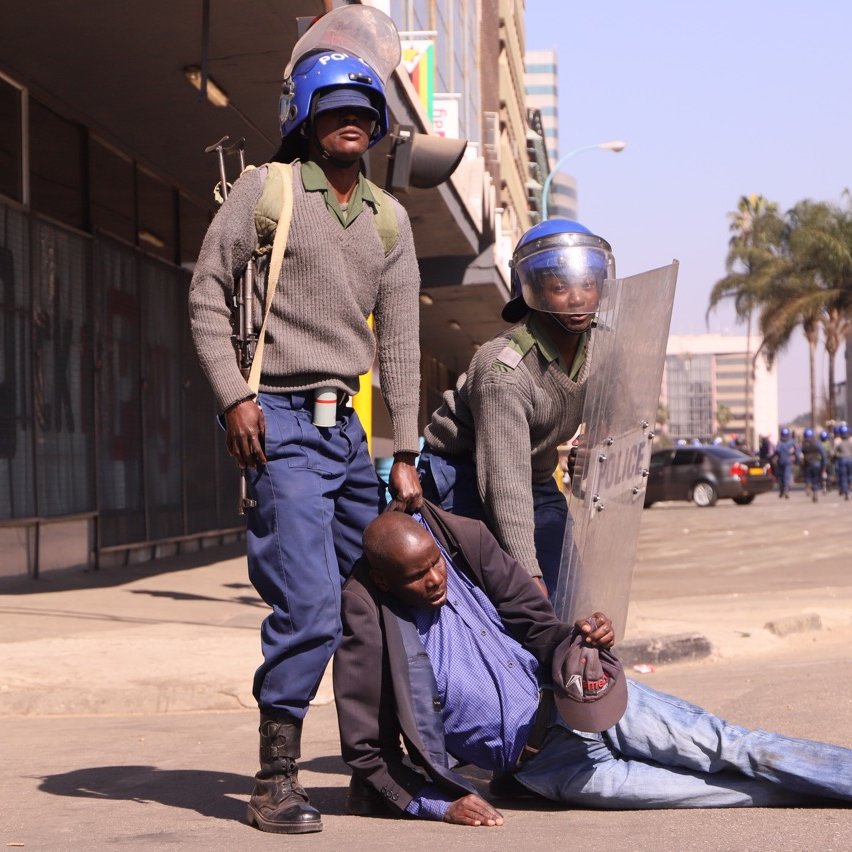 L'image montre une scène de tension dans un environnement urbain. Deux policiers, en uniforme et portant des casques de protection, semblent maîtriser un homme qui est au sol. L'homme est vêtu d'une chemise foncée et d'une veste. Il semble être en difficulté, soutenu par l'un des policiers, tandis que l'autre se tient à ses côtés avec un bouclier. En arrière-plan, on peut apercevoir des bâtiments modernes et quelques palmiers, indiquant que la scène se déroule dans une ville. L'atmosphère générale semble tendue, évoquant un moment de confrontation.