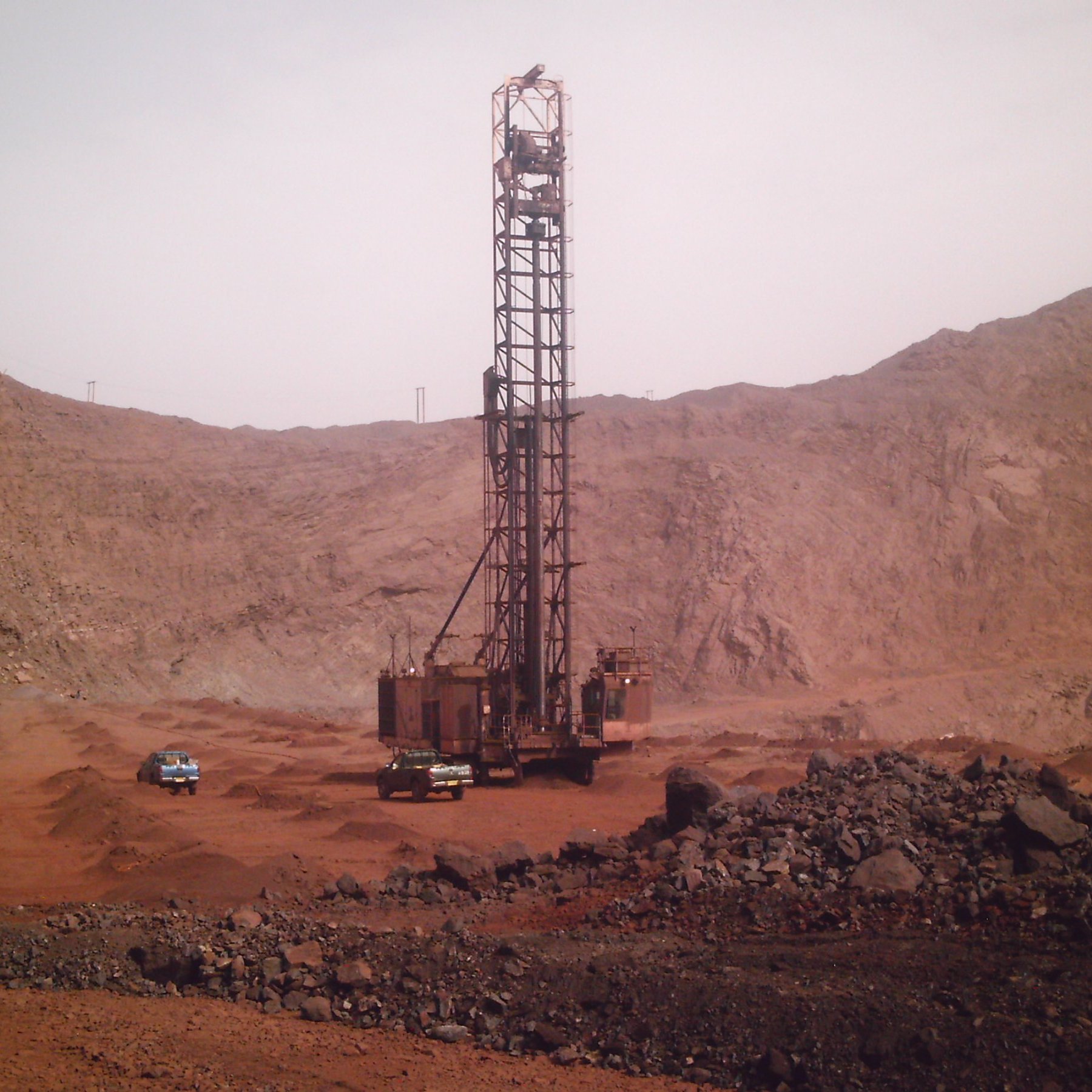 L'image montre un site d'exploitation minière dans un environnement aride et rocheux. Au centre, une grande foreuse se dresse, entourée de montagnes de terre et de roches. La foreuse, de couleur rouille, a une structure métallique imposante qui semble clé dans le processus d'extraction. Autour d'elle, plusieurs véhicules, probablement des camions ou des 4x4, sont garés sur le sol. Le terrain environnant est rocailleux et dépouillé, avec des teintes ocres et brunes, évoquant une atmosphère sévère et industrielle. Le ciel est clair, ce qui accentue l'impression de chaleur et d'aridité de cette zone minière.