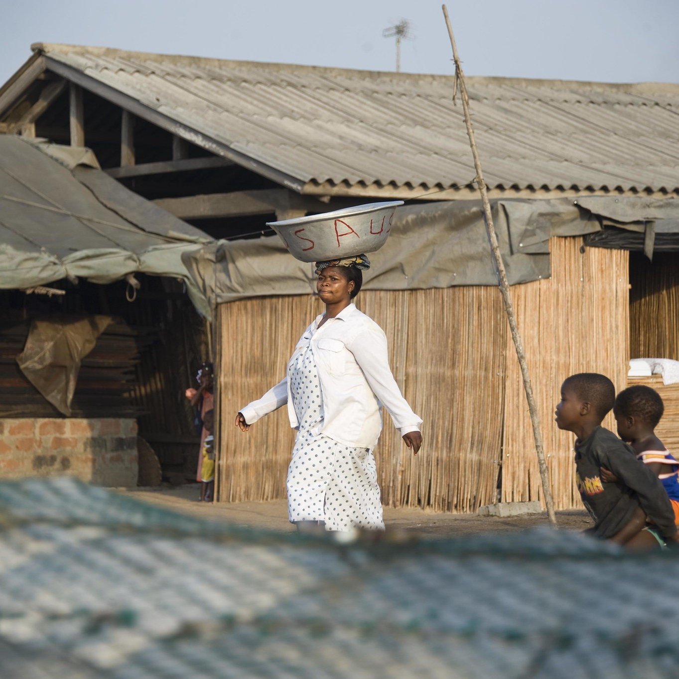 Dans cette scène vibrante, une femme porte une grande bassine sur sa tête, symbole de son travail quotidien. Elle avance avec assurance sur un chemin, tout en étant entourée de maisons à architecture simple, en bois et en bambou. Le ciel est clair, laissant présager une belle journée ensoleillée. À proximité, des enfants jouent ensemble, ajoutant une atmosphère de vie et de dynamisme à l'environnement. Les ombres et les textures des matériaux environnants se mêlent, créant un paysage riche et vivant. L'ensemble dégage une ambiance d'activité et de communauté.