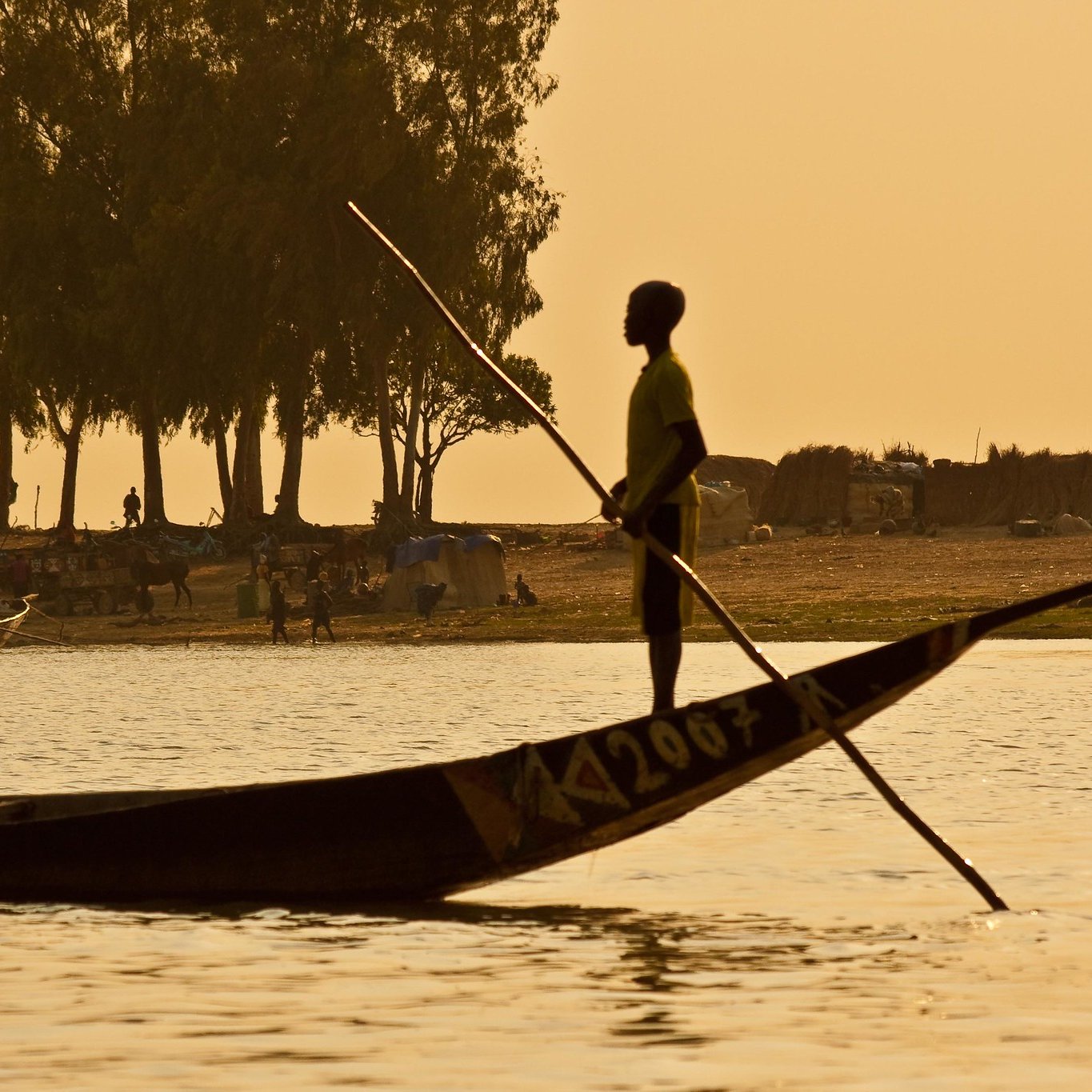 Imaginez une scène paisible au bord d'un fleuve. Un jeune garçon se tient debout dans une pirogue traditionnelle, les pieds ancrés sur le fond de la barque. Il utilise une longue perche pour diriger son embarcation à travers l'eau calme. En arrière-plan, on peut apercevoir un ruisseau de silhouettes d'arbres élancés, probablement des manguiers ou des sycomores, se découpant contre le ciel orangé du crépuscule. Sur la rive, des groupes de personnes s'affairent, tandis que des abris faits de toile et de paille sont visibles, suggérant une vie communautaire active. L'atmosphère est tranquille, teintée d'une chaleur douce, et l'ensemble dégage une sensation de sérénité et de connexion à la nature.