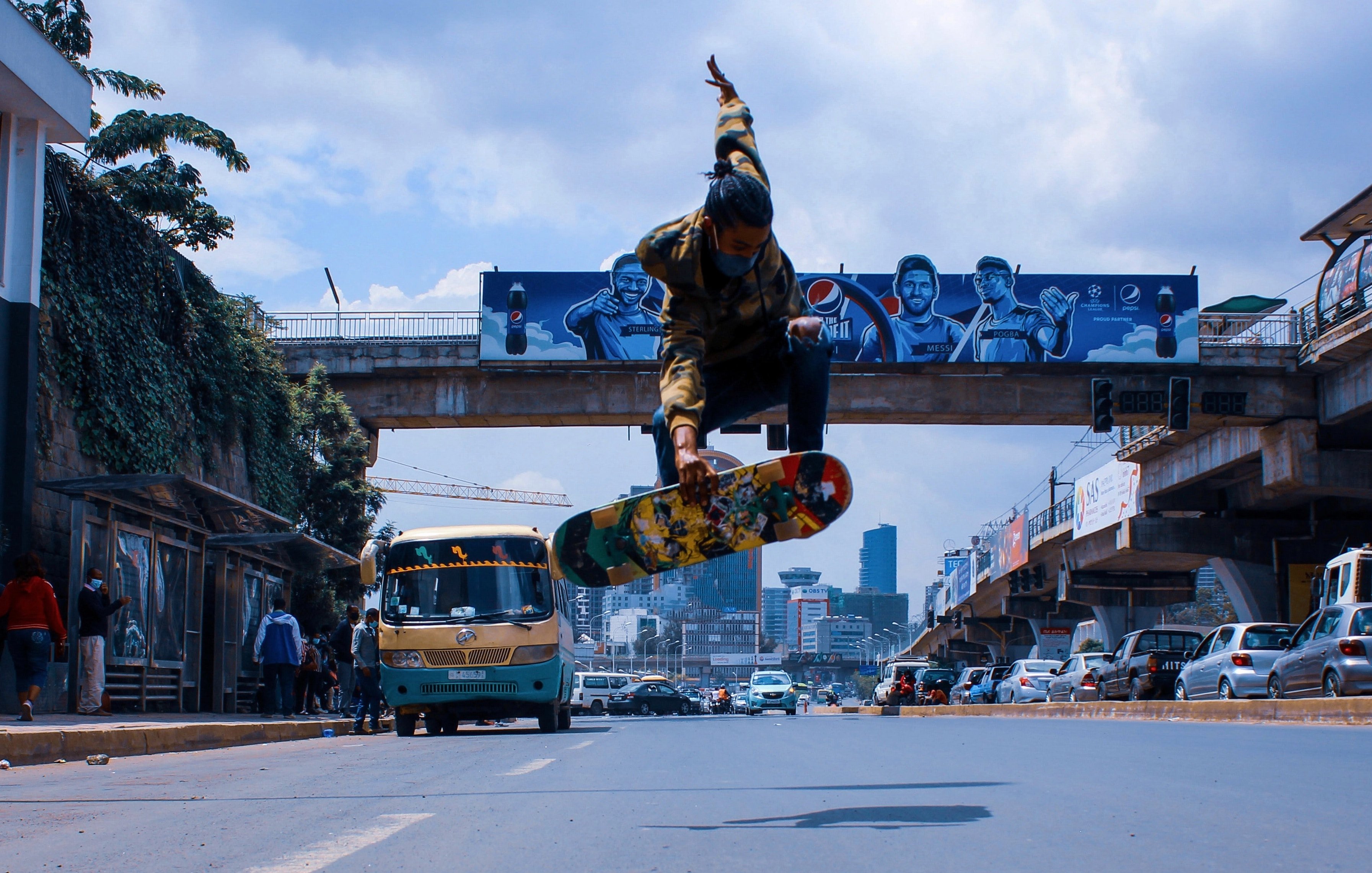 L'image montre un skateur en plein saut au-dessus de la route. Il est habillé d'un sweat à capuche avec des motifs et porte un skateboard coloré. En arrière-plan, il y a des bâtiments urbains, des graffitis sur un mur, et un bus qui passe, typique des grandes villes. La scène capture une ambiance dynamique et énergique, avec un ciel partiellement nuageux qui ajoute à l'atmosphère urbaine. Les paysages urbains et la présence d'autres personnes montrent l'animation de la ville.