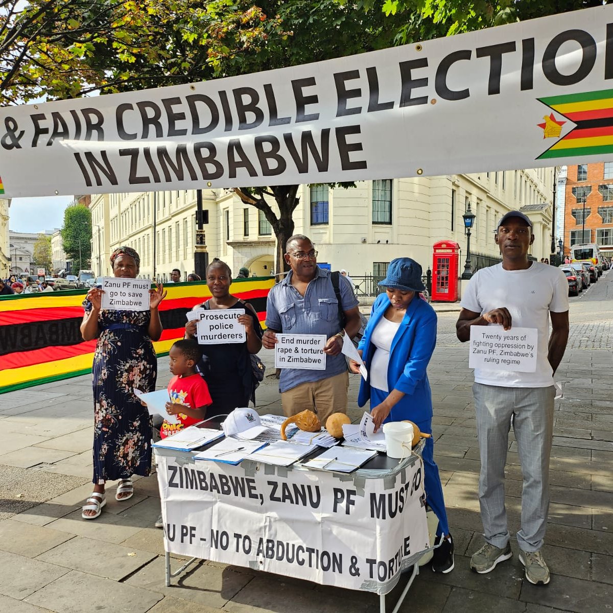 L'image montre un groupe de personnes rassemblées sous une grande banderole qui proclame "Élections libres et équitables au Zimbabwe". Cette banderole est ornée des couleurs du drapeau zimbabwéen, avec du rouge, du jaune, du vert et du noir. Les manifestants tiennent des panneaux avec des messages portant sur la nécessité de respecter les droits humains et d'organiser des élections justes au Zimbabwe. Sur la table devant eux, il y a des brochures et des documents qui semblent orientés vers la sensibilisation et l'information sur la situation politique. L'environnement est urbain, avec des bâtiments en arrière-plan et des passants qui s'intéressent à la manifestation.