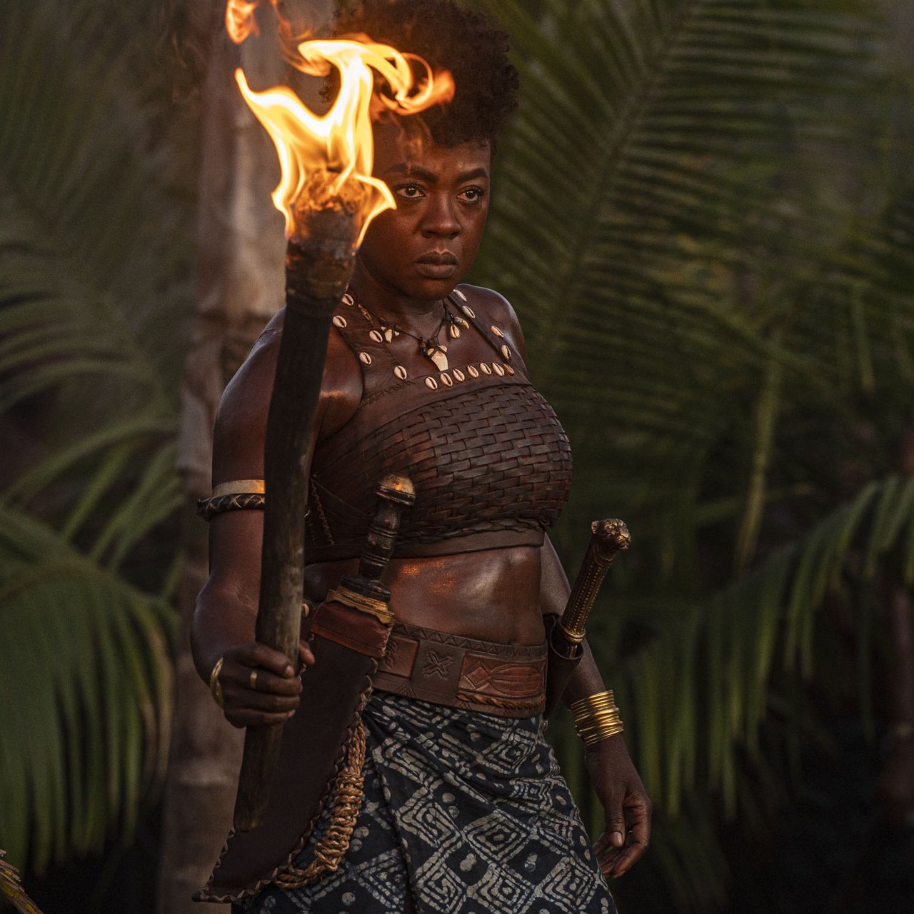 L'image montre une femme debout au milieu d'une luxuriante végétation tropicale, avec des palmiers en arrière-plan. Elle tient une torche enflammée dans sa main droite, créant une lumière chaleureuse qui éclaire son visage déterminé. Elle porte un haut traditionnel en cuir et une jupe décorée de motifs. Des bracelets dorés ornent ses bras, ajoutant une touche d'élégance à son apparence. Son expression est forte et intense, reflétant à la fois courage et détermination. L'atmosphère de l'image évoque une ambiance de puissance et de leadership.