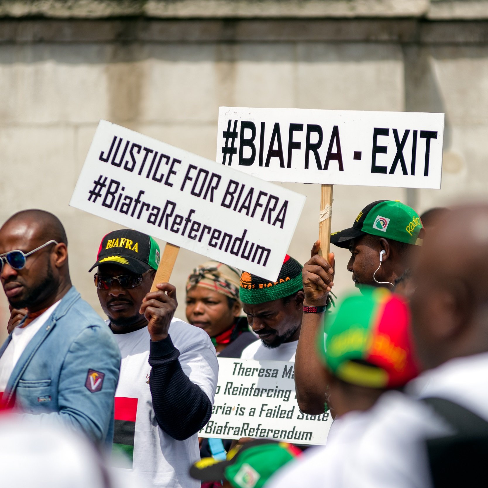 L'image montre un groupe de personnes participant à une manifestation. Elles portent des t-shirts blancs et des casquettes aux couleurs du mouvement Biafra. Des pancartes sont brandies, portant des inscriptions telles que "#BIAFRA - EXIT" et "JUSTICE FOR BIAFRA", ainsi que des hashtags liés au référendum biafrais. L'atmosphère semble dynamique et engagée, reflétant une revendication pour la justice et l'autodétermination.