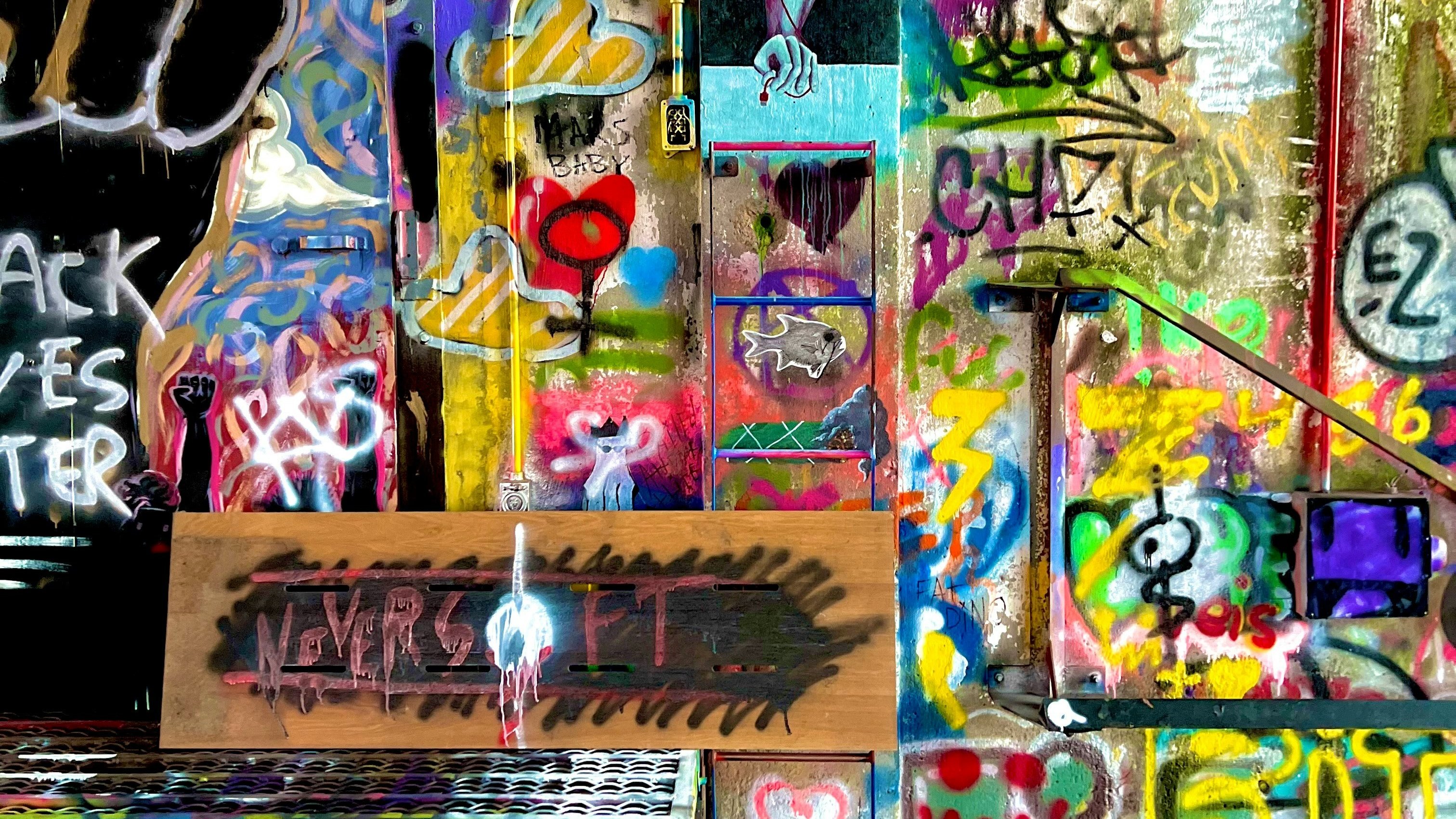 Cette image montre un mur vibrant de graffiti et d'art urbain. Les couleurs éclatantes, comme le jaune, le rouge, le bleu et le vert, créent un tableau chaotique et dynamique. On peut y voir des symboles et des mots, notamment un poing levé, représentant la résistance, ainsi que des cœurs et des dessins abstraits. Une planche en bois au centre affiche des mots en noir, évoquant des thèmes de persévérance. L'ensemble dégage une atmosphère de créativité libre et d'expression personnelle, transformant un espace ordinaire en une œuvre d'art collective.