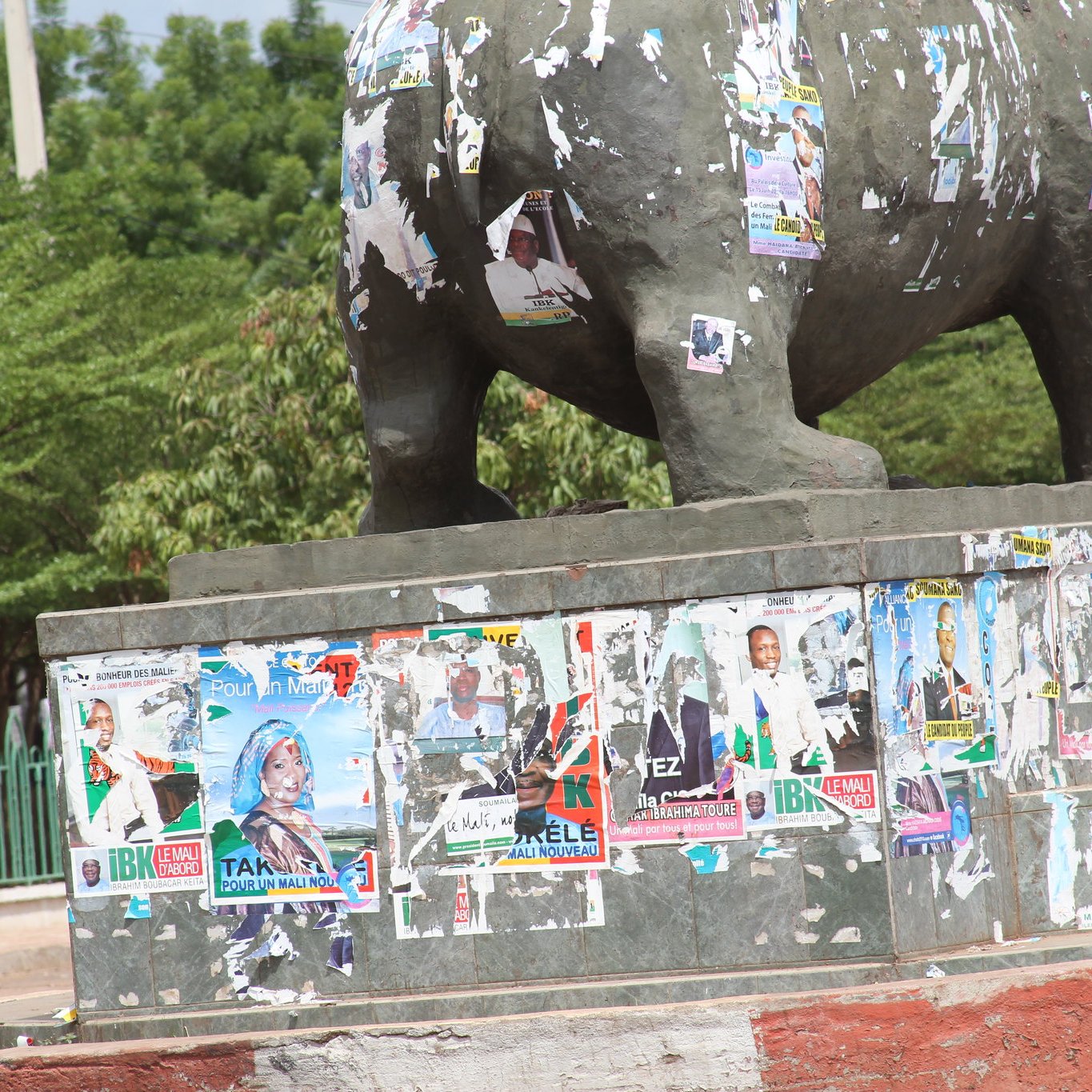 L'image montre une grande statue d'éléphant, probablement en bronze ou en métal, se tenant sur un socle en béton. La statue est recouverte de nombreuses affiches publicitaires et politiques, dont certaines sont déchirées et se détachent. Ces affiches présentent des visages et des textes, indiquant des campagnes électorales, des événements ou des promotions. Le socle en béton est partiellement peint en rouge et en gris, ajoutant un contraste à l'image. À l'arrière-plan, on aperçoit des arbres verdoyants et d'autres éléments de la rue, ce qui suggère une ambiance vivante et urbaine. Les couleurs sont variées et l'aspect général est un mélange de textures et de visuels, témoignant d'une interaction entre la nature et l'urbanisme.