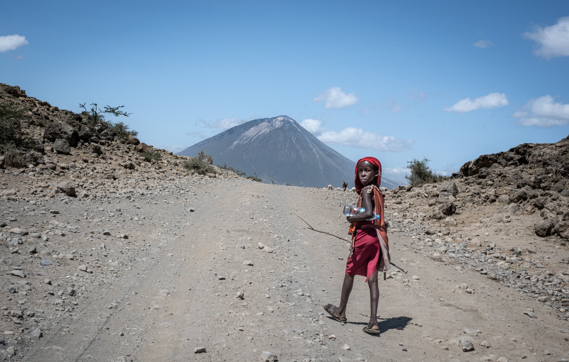 L'image montre un enfant marchant sur un chemin de terre. Il porte une robe rouge et un foulard, et il semble porter un petit récipient ou une bouteille dans ses mains. En arrière-plan, on aperçoit un volcan majestueux, dont le sommet est légèrement nuageux sous un ciel bleu clair parsemé de quelques nuages blancs. Le terrain autour est rocailleux et aride, avec quelques petits buissons éparpillés. L'ensemble de la scène dégage une atmosphère de nature sauvage et éloignée.