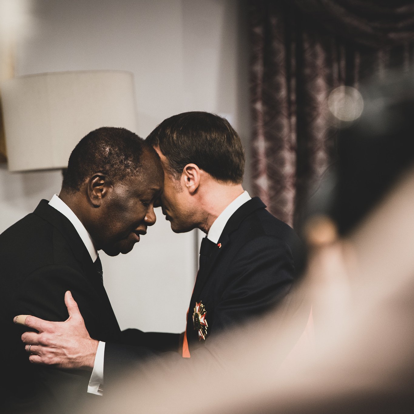 L'image montre deux hommes en train de se saluer de manière chaleureuse, leurs fronts se touchant, ce qui suggère une connexion profonde ou un moment d'émotion. L'un des hommes porte un costume sombre, tandis que l'autre semble également en habit formel. Le décor en arrière-plan, légèrement flou, indique un environnement intérieur, probablement un bureau ou une salle de conférences. La lumière est douce et crée une atmosphère respectueuse et intime. Les expressions des deux hommes semblent sérieuses et significatives, évoquant un échange important ou une reconnaissance mutuelle.