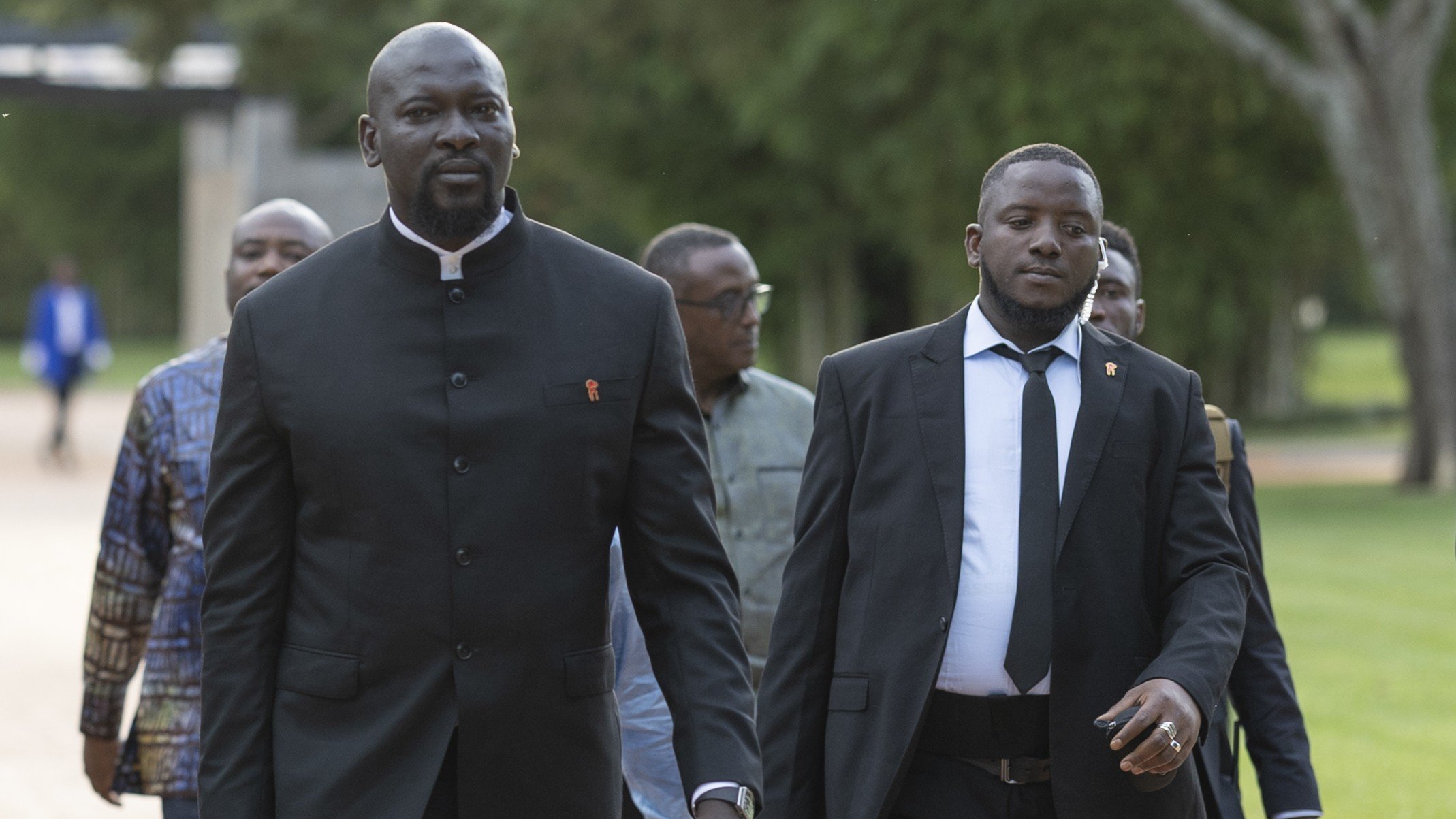 The image depicts a group of several men walking together in a park-like setting. The man in the foreground, dressed in a black suit with a high collar, appears confident and focused. He is accompanied by another man wearing a black suit and tie, who carries a briefcase. The background features lush greenery, suggesting an outdoor event or gathering. Other men can be seen following closely behind, contributing to a formal yet relaxed atmosphere.