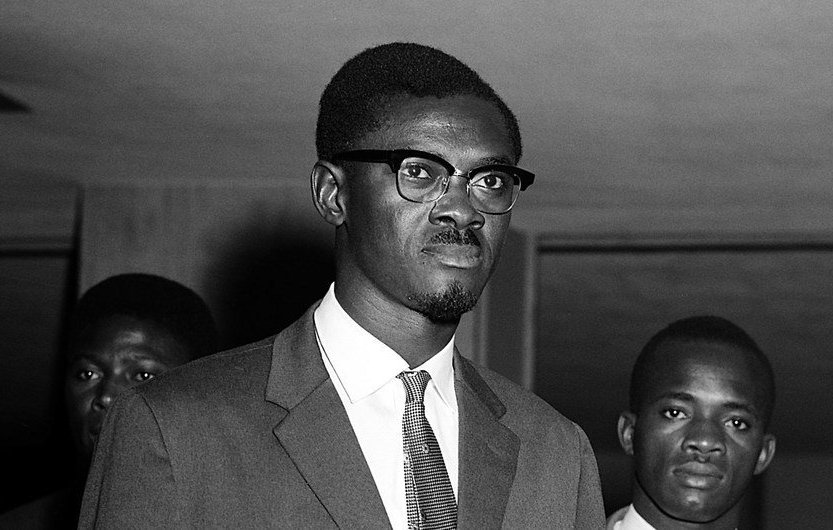 L'image montre un groupe d'hommes en costume, pris dans un cadre formel. Au premier plan, un jeune homme porte des lunettes à monture noire et affiche une expression sérieuse. Il est vêtu d'un costume sombre avec une chemise claire et une cravate. Derrière lui, deux autres hommes sont présents, également en costume, portant des expressions neutres. La lumière est douce, et l'atmosphère dégage un sentiment de gravité ou d'importance. Les murs autour d'eux semblent être peu décorés, mettant l'accent sur les personnes au centre de l'image.