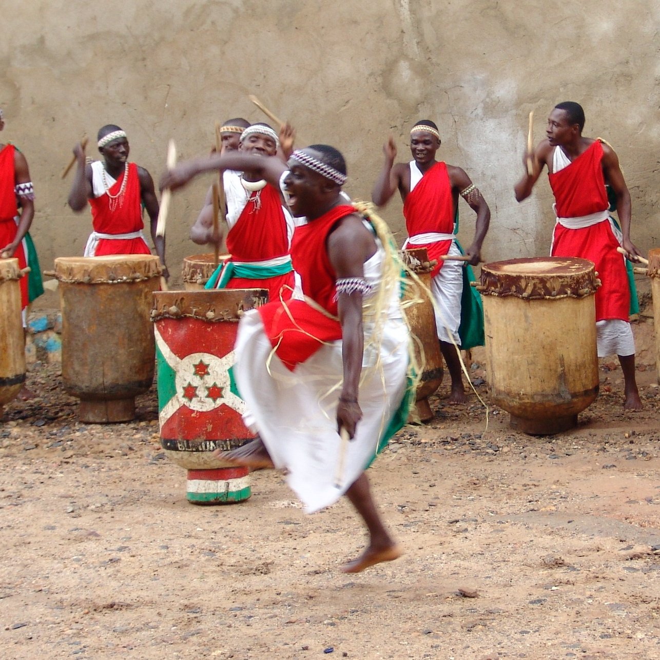 L'image représente un groupe de percussionnistes en pleine performance. Ils portent des vêtements traditionnels, principalement rouges, avec des ceintures vertes. Au centre, un homme se distingue en sautant avec énergie, créant un mouvement dynamique. Les autres membres du groupe, en arrière-plan, jouent des tambours en bois de forme cylindrique. L'atmosphère est festive et rythmée, avec une ambiance de célébration culturelle, où les sons des tambours résonnent fortement. Le sol est en terre, ce qui ajoute un aspect rustique à la scène.