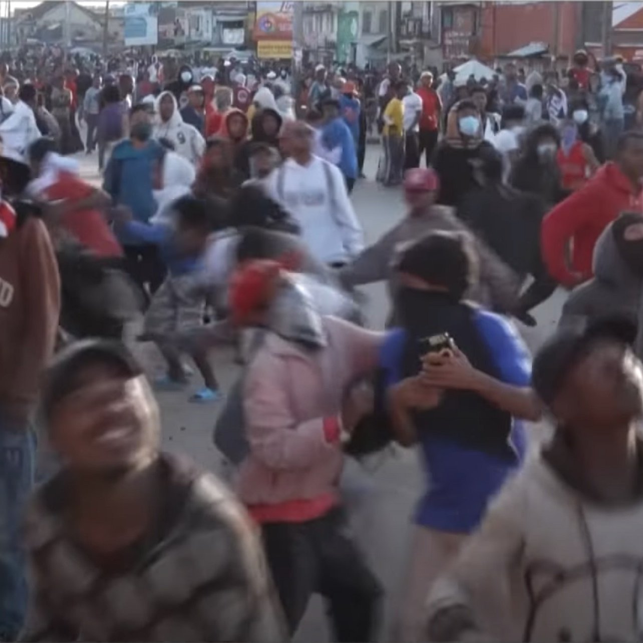 L'image montre une foule dense de personnes rassemblées dans une rue, probablement lors d'une manifestation ou d'un événement populaire. Les participants portent des vêtements variés et beaucoup d'entre eux ont des masques ou des foulards sur le visage. On peut sentir une ambiance animée et énergique, avec des mouvements rapides et des gestes expressifs. Certains individus semblent prendre des photos avec leurs téléphones. L'arrière-plan est rempli d'autres personnes, créant une atmosphère vibrante et collective.