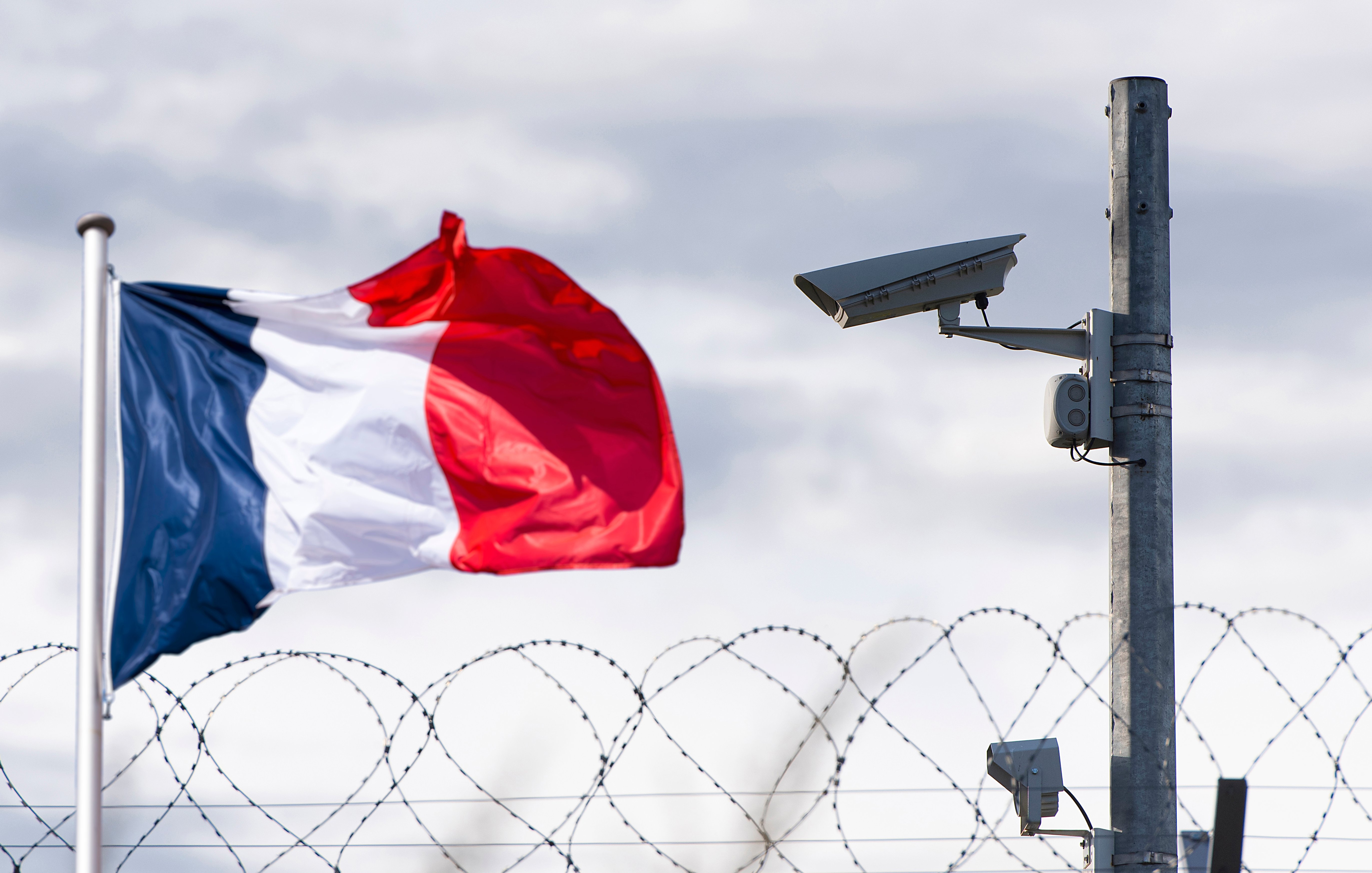 L'image montre un drapeau français vibrant, flottant au vent. À côté, on aperçoit une caméra de surveillance fixée sur un poteau. En arrière-plan, un fil de fer barbelé accentue l'idée de sécurité et de surveillance. Le ciel est nuageux, ajoutant une atmosphère à la scène, qui évoque un environnement protégé.