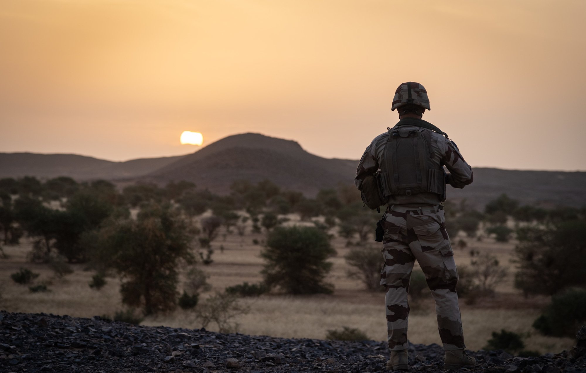 La scène se déroule dans un paysage désertique au coucher du soleil. Au premier plan, un soldat en uniforme camouflage se tient debout, tourné vers l'horizon. Sa silhouette est mise en valeur par la lumière dorée du soleil qui descend lentement derrière une colline arrondie. Le terrain est rocailleux et parsemé de petites végétations et arbres, typiques des paysages arides. L'ambiance est calme, presque contemplative, ajoutant une dimension de réflexion à la présence du soldat, probablement en mission. Les couleurs chaudes du ciel se mélangent à la beauté naturelle du paysage, créant une atmosphère à la fois sereine et intense.