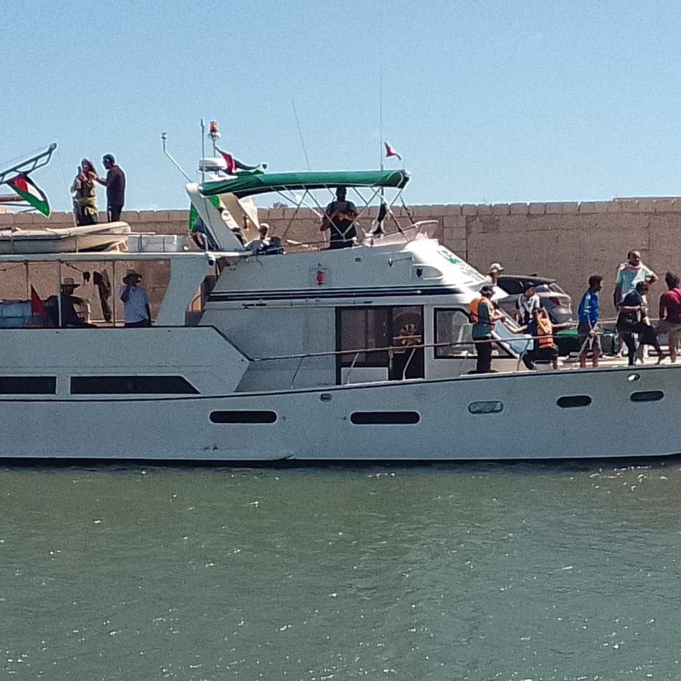 Imaginez un bateau de plaisance élégant, amarré à un quai au bord de l'eau. Sa coque est blanche, et le soleil illumine son pont supérieur qui est équipé d'un auvent vert. Plusieurs personnes se trouvent sur le bateau et sur le quai ; certaines semblent s'apprêter à embarquer, tandis que d'autres discutent joyeusement. L'arrière-plan montre un mur de pierre beige qui contraste avec le bleu de l'eau. L'atmosphère est animée et estivale, évoquant une journée de détente en bord de mer.