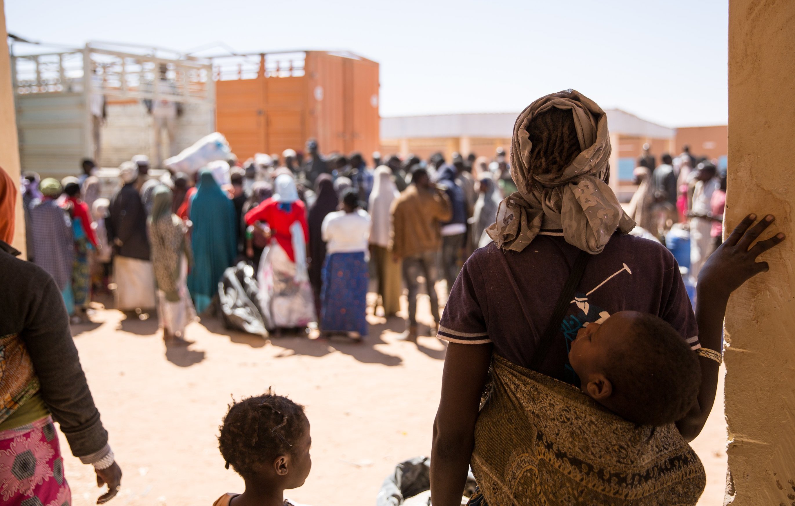 L'image montre une scène poignante dans un contexte de rassemblement de personnes, probablement dans un camp ou un centre d'accueil. Au premier plan, une personne, probablement une mère, porte un enfant dans un sling. Elle regarde vers un groupe de personnes, qui attendent patiemment, formant une masse compact de silhouettes. La majorité des personnes dans le fond sont habillées de vêtements aux couleurs variées, certaines portant des foulards ou des vêtements traditionnels. Le sol est en terre battue, et des containers peuvent être aperçus à l'arrière-plan, suggérant un environnement temporaire. L'atmosphère semble à la fois calme et chargée d'une certaine tension, reflétant une réalité complexe.