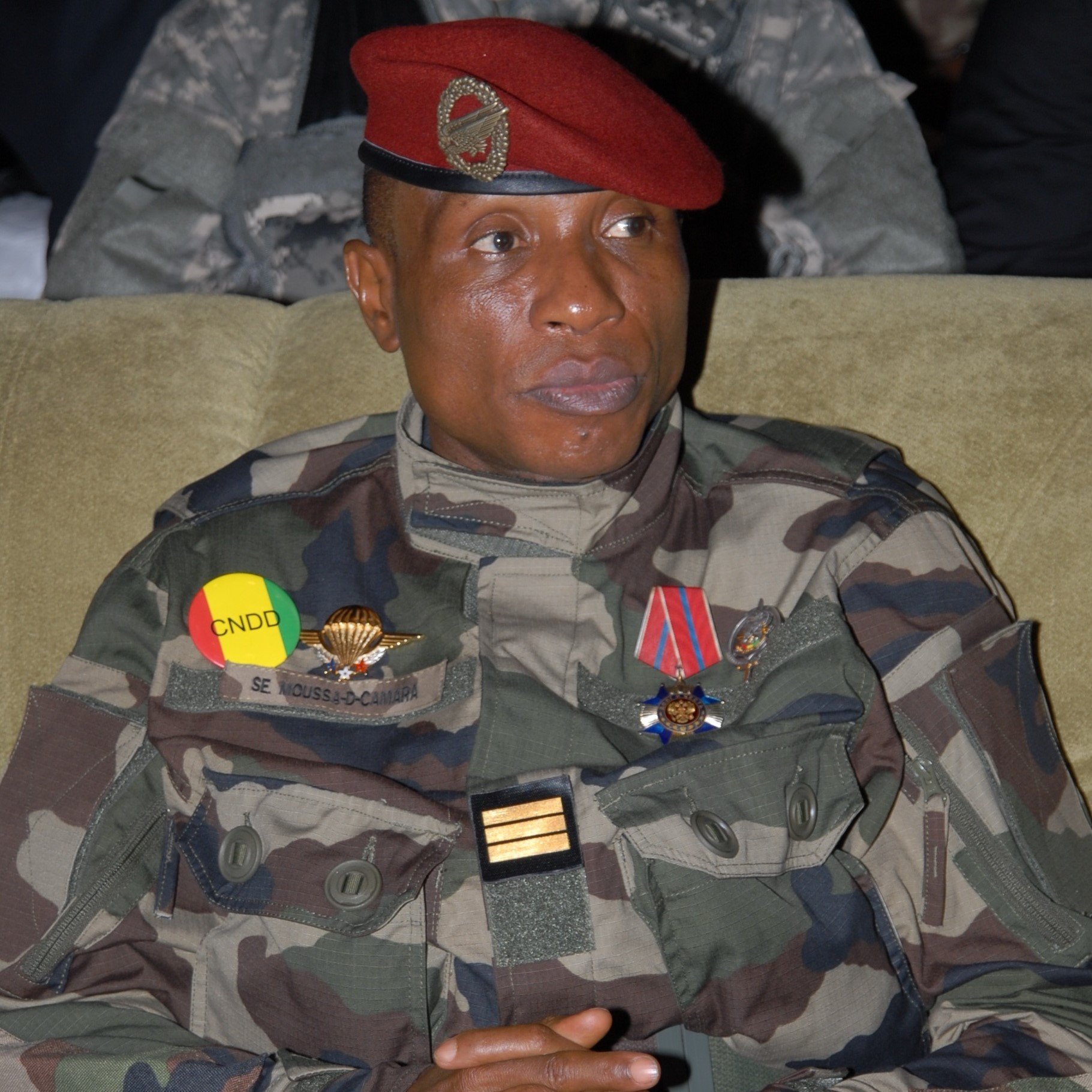 L'image montre un militaire assis, vêtu d'un uniforme camouflage. Il porte une casquette rouge ornée d'un insigne. Sur sa poitrine, il y a plusieurs décorations et médailles visibles. Les couleurs de l'uniforme et des insignes, notamment le jaune et le rouge, évoquent des éléments de son pays. Son expression est sérieuse, et il semble attentif, probablement à une discussion ou à un événement. L'arrière-plan est flou, suggérant une atmosphère formelle ou solennelle.