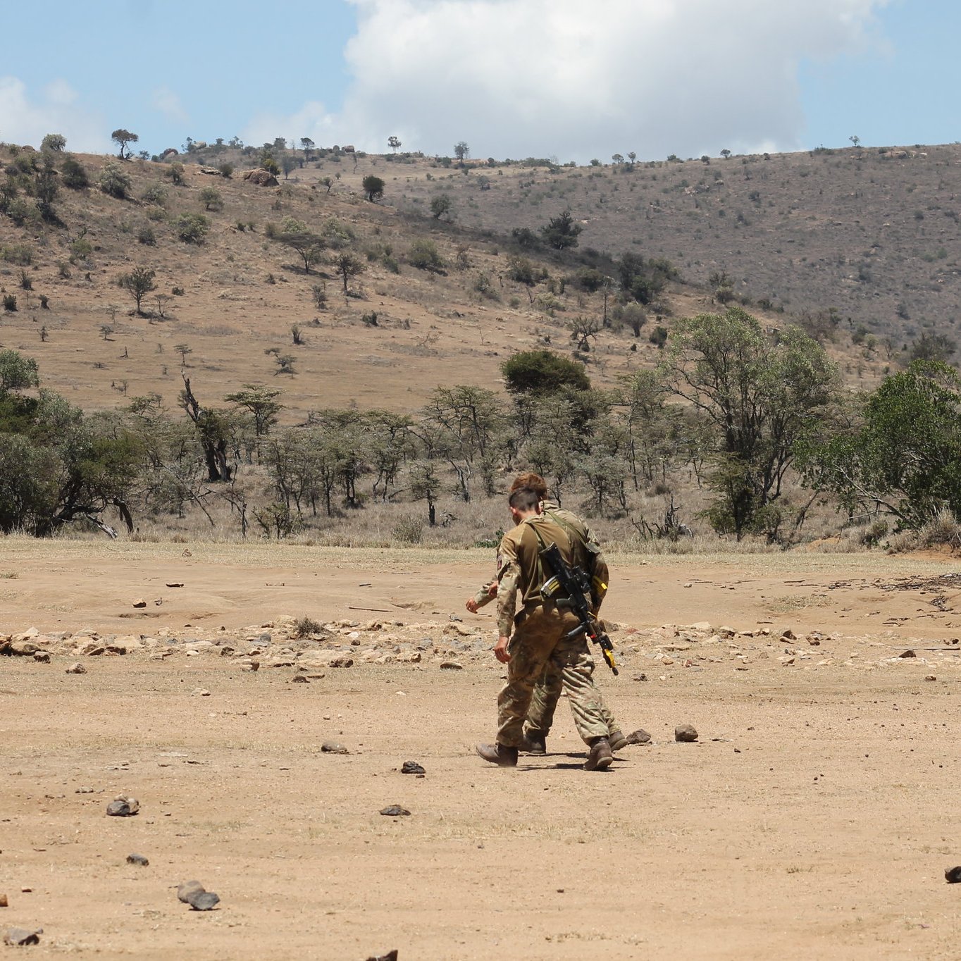 L'image montre un paysage aride et vaste, typique de la savane. Au centre, un homme marche seul sur un sol poussiéreux, parsemé de pierres. Il est vêtu d'un uniforme camouflage et porte un équipement à son dos, suggérant une activité en milieu naturel. En arrière-plan, on aperçoit des collines sèches et quelques arbres épars, témoignant d'une végétation clairsemée. Le ciel est partiellement nuageux, avec des nuages blancs qui contrastent avec le bleu du ciel. L'ensemble dégage une atmosphère de solitude et d'exploration dans un environnement sauvage.