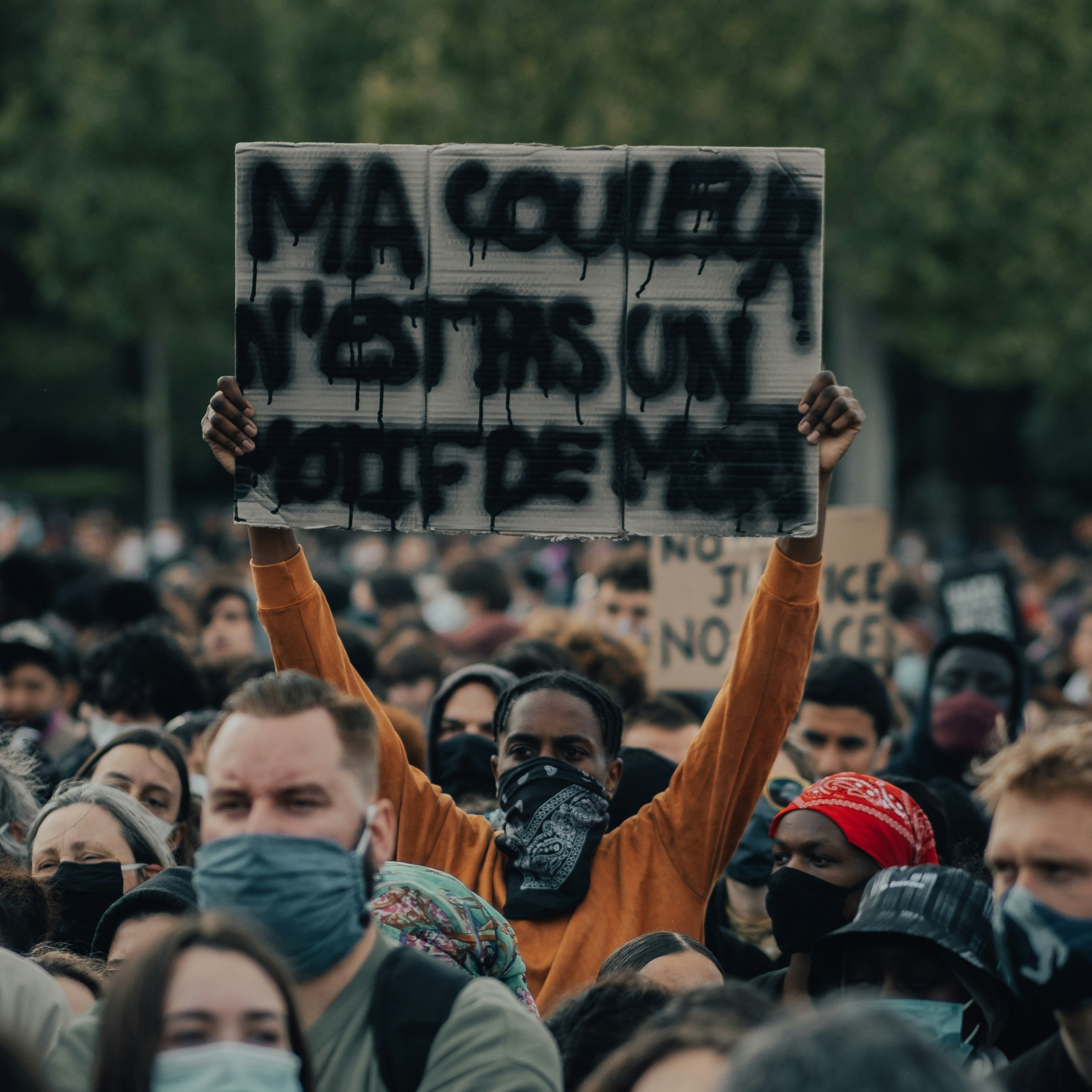 L'image montre une grande foule rassemblée pour une manifestation. Parmi les manifestants, une personne se distingue en tenant une pancarte sur laquelle est écrit "MA COULEUR N'EST PAS UN CRIME". Cette personne porte un masque, comme beaucoup d'autres dans la foule. Les visages autour sont sérieux et déterminés, reflétant un sentiment de solidarité et de revendication. On peut sentir l'intensité de l'événement grâce à l'énergie collective. En arrière-plan, des arbres indiquent un cadre naturel, ajoutant à l'atmosphère de mobilisation pacifique.