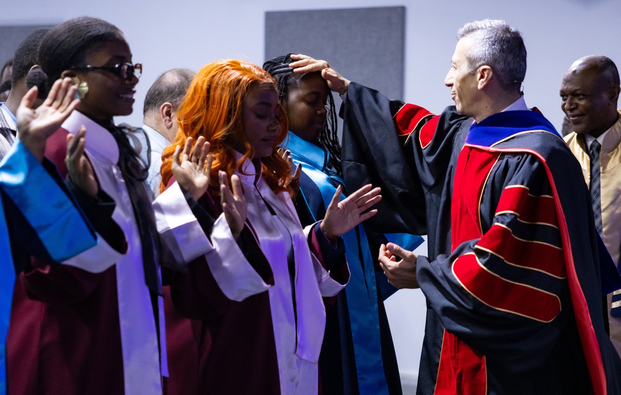 L'image montre une scène de cérémonie de remise des diplômes dans un auditorium. Plusieurs diplômés sont présents, portant des toges de différentes couleurs : certaines en bordeaux et d'autres en bleu. Ils sont debout en ligne, avec des expressions qui expriment à la fois la fierté et l'anticipation. Un homme, vraisemblablement un responsable de l'établissement, se tient devant eux, en train de poser une main sur la tête d'une diplômée avec des cheveux bouclés et roux. Les autres diplômés lèvent les mains, soit pour montrer leur gratitude, soit en signe de célébration. En arrière-plan, des personnes supplémentaires assistent à l'événement, complétant l'atmosphère festive de cette occasion mémorable.
