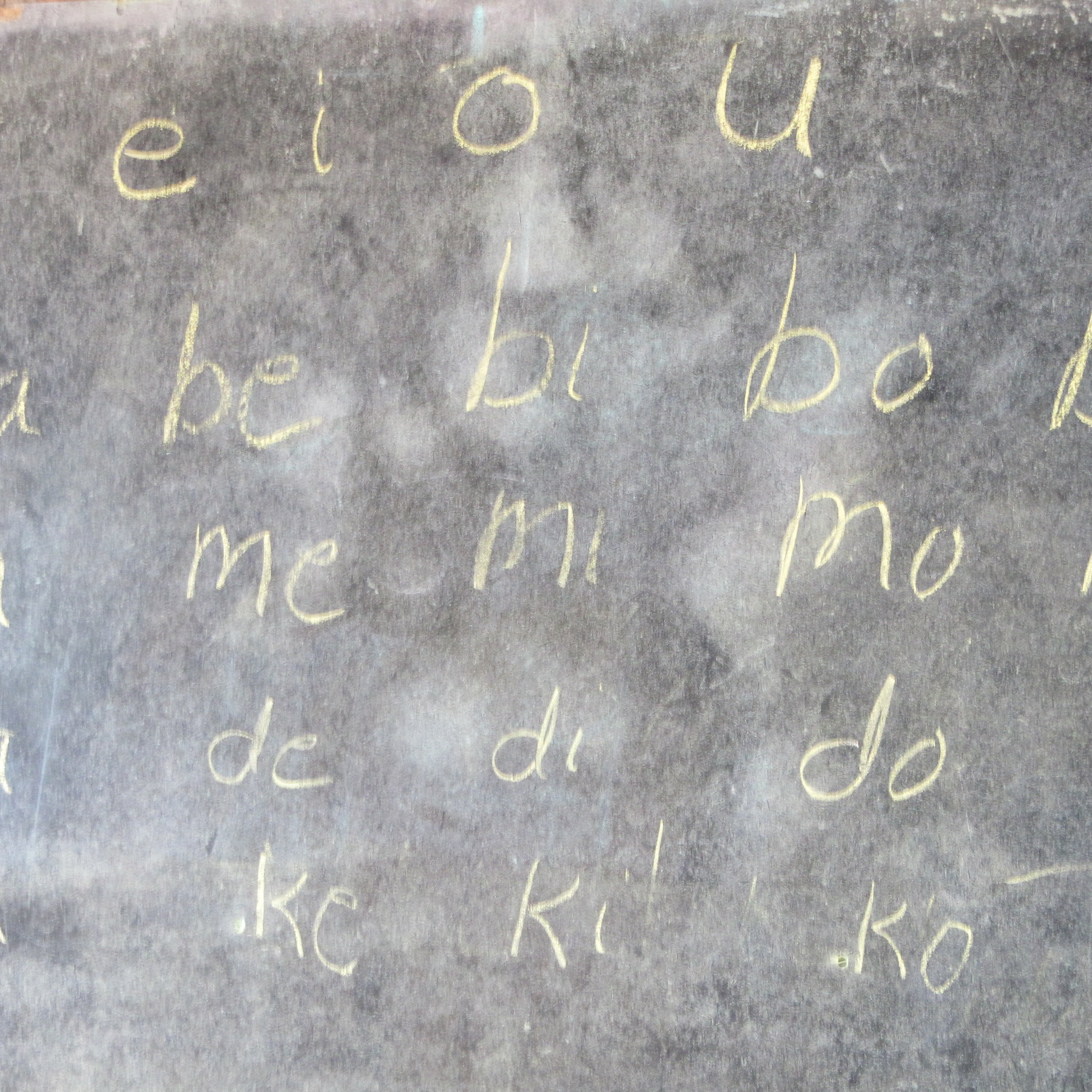 L'image montre un tableau noir sur lequel sont écrits des caractères en lettres cursives, à la craie. Les lignes commencent par les voyelles "a", "e", "i", "o", "u", suivies de combinaisons de consonnes et voyelles, telles que "ba", "be", "bi", "bo", "bu", puis "ma", "me", "mi", "mo", "mu", et ainsi de suite. L'écriture est claire et simple, et le tableau a une texture légèrement rugueuse. L'ensemble de l'image évoque un environnement d'apprentissage.