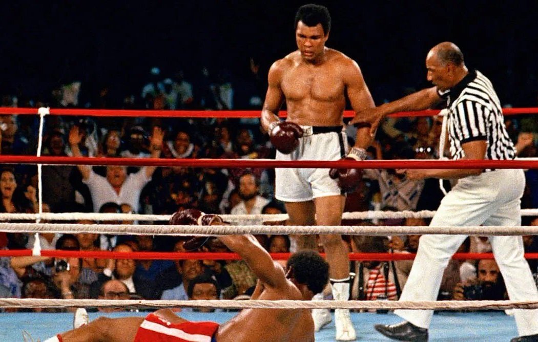 L'image montre un moment mémorable d'un match de boxe. Au centre, un boxeur, torse nu et portant des shorts blancs, se tient debout avec un regard déterminé. C'est Muhammad Ali, connu pour sa rapidité et sa technique. À ses pieds, un autre boxeur est allongé sur le sol, faisant face au ring, après avoir été mis au tapis. Le public en arrière-plan est en émoi, certains levant les bras en signe d'enthousiasme ou d'étonnement. Un arbitre, vêtu d'un maillot à rayures, s'approche du boxeur au sol, prêt à commencer le compte. L'atmosphère est chargée d'intensité et d'adrénaline, typique des combats de boxe.
