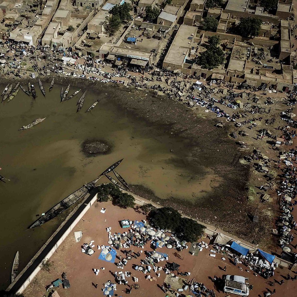 Imaginez un paysage vu du ciel. Au centre, un plan d'eau sombre qui semble être une lagune ou un port. Les bords de l'eau sont bordés de bateaux, certains amarrés, d'autres flottant paisiblement. Autour de cette lagune, il y a des habitations, des maisons en terre ou en tuiles, formant un petit village. Sur les rives, des gens sont rassemblés, certains assis par terre, d'autres se déplaçant avec des marchandises. Des tentes et des parasols sont disposés, suggérant un marché vivant où l'on troque et vend des produits. L'air est probablement rempli de sons, de cris et d'odeurs de nourriture, de poissons et de marchandises variées, donnant l'impression d'une scène animée et prospère. Les couleurs sont chaudes, évoquant un climat ensoleillé. Ce tableau dégage une atmosphère d'activité et de communauté autour de l'eau.