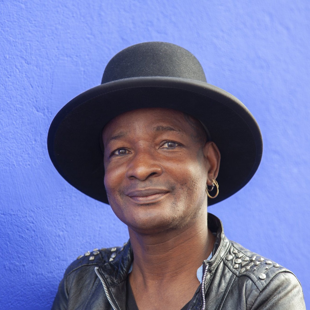 Cette image montre un homme souriant, portant un grand chapeau noir qui a une forme de haut de forme. Il est habillé d'une veste en cuir noire ornée de clous, ce qui lui donne un style à la fois décontracté et audacieux. L'arrière-plan est d'un bleu vif, créant un contraste frappant avec ses traits. Son expression est amicale et accueillante, évoquant une personnalité chaleureuse et charismatique.