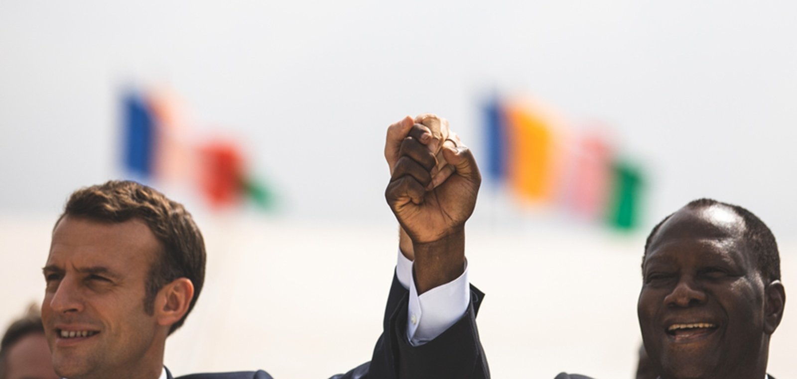 The image shows two men, one wearing a suit with a tie, raising their hands together in a gesture of solidarity or celebration. They are smiling and appear to be in a public setting. In the background, there are several flags visible, suggesting an international context or event. The atmosphere seems positive and hopeful.