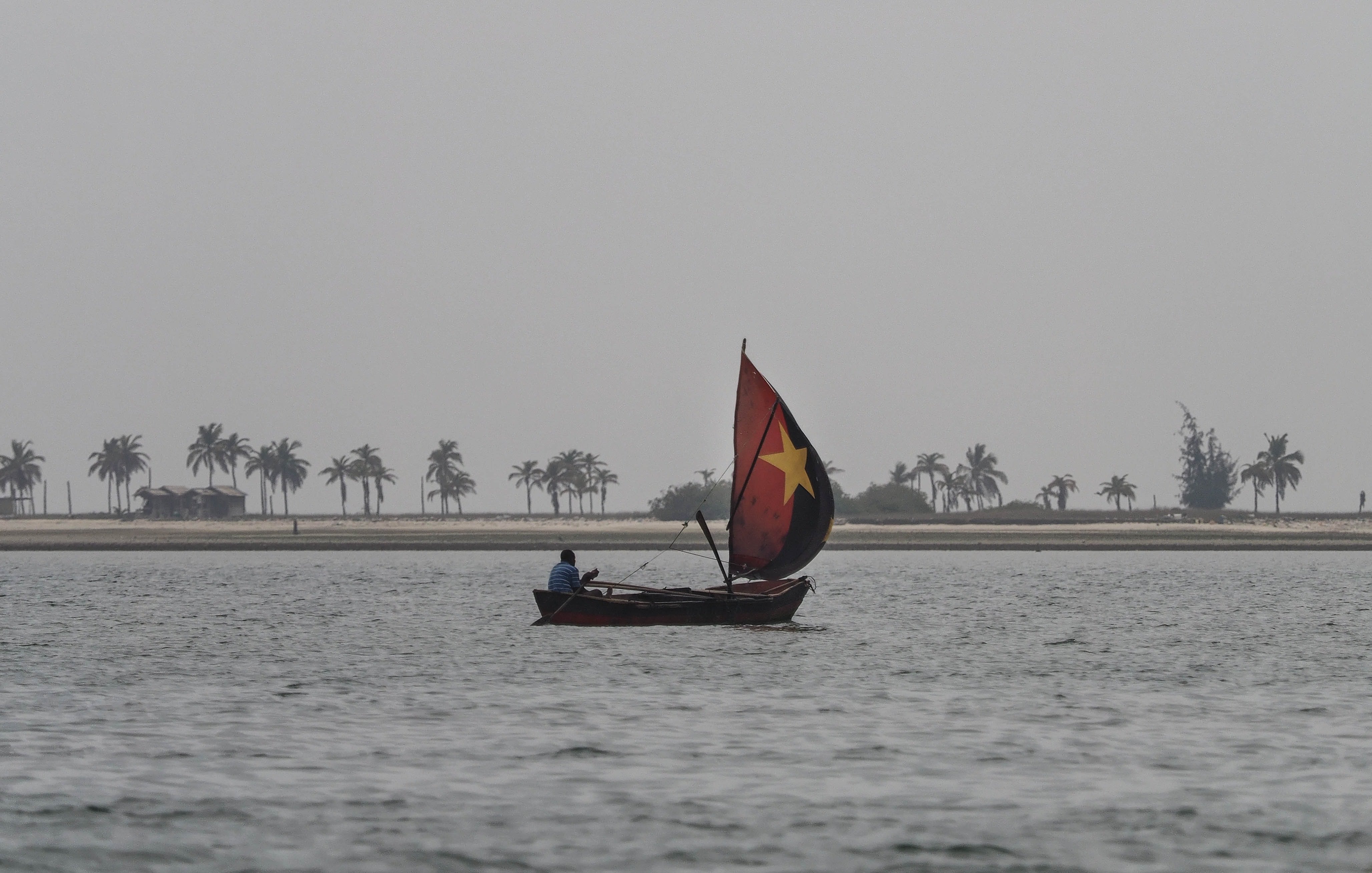 L'image montre une scène calme sur l'eau, où un petit bateau à voile glisse doucement sur des vagues légères. Le voilier est décoré d'un grand drapeau aux couleurs vives, incluant une étoile, qui se distingue dans le paysage. Au loin, on aperçoit une plage bordée de palmiers qui se dressent contre un ciel nuageux et brumeux. L'atmosphère est paisible, suggérant un moment de solitude et de tranquillité au bord de l'eau. La douceur du vent et l'écho des vagues contribuent à créer une ambiance sereine.
