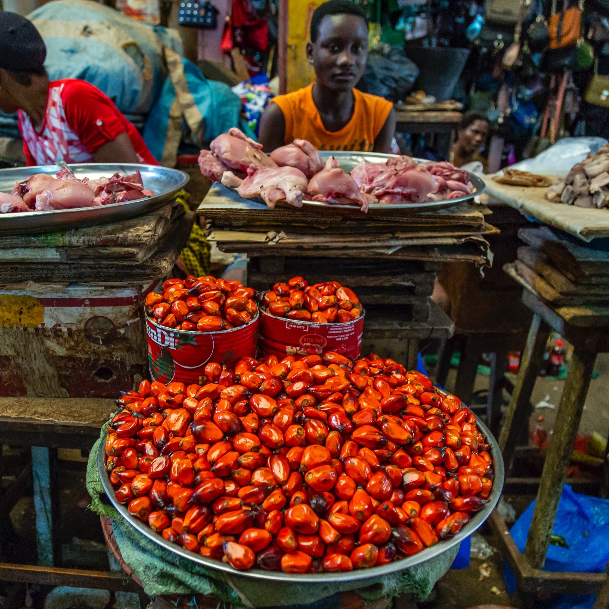 L'image montre un marché animé, où des étals en bois sont chargés de différents aliments. Au premier plan, un grand plat en métal contient des petites noix rouge vif, à côté d'autres aliments. À l'arrière-plan, des morceaux de viande sont disposés sur des plateaux, et on peut voir des vendeurs et des clients s'affairer autour. Les couleurs sont vives, avec des nuances de rouge, de marron et de vert. L'atmosphère est dynamique, remplie de sons de discussions et d'échanges.