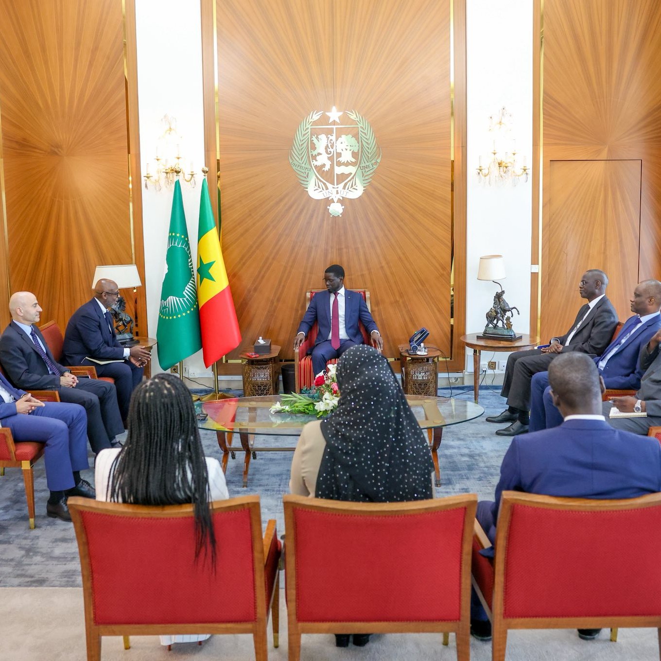 L'image montre une réunion dans une salle élégante. Au centre, un homme assis porte un costume et semble être à la tête de la discussion. Autour de lui, plusieurs personnes assisent dans des fauteuils confortables. À l'arrière-plan, on peut voir les drapeaux du Cameroun et du Sénégal. La pièce est lumineuse, avec des rideaux drapés et un décor raffiné, donnant une atmosphère formelle et professionnelle à cette rencontre. Les participants semblent engagés dans une conversation sérieuse.