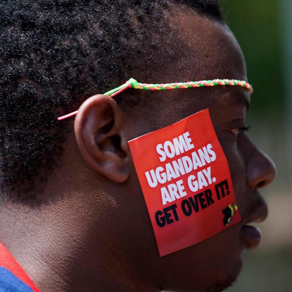 L'image représente un homme dont le visage est de profil. Il porte un petit autocollant sur sa joue qui affiche un message en lettres blanches sur fond rouge. Le texte dit : "SOME UGANDEANS ARE GAY. GET OVER IT!" (Certains Ougandais sont gays. Acceptez-le !). L'homme a des cheveux courts et un accessoire coloré autour de la tête. L'ambiance générale de l'image suggère un acte de protestation ou de revendication des droits.
