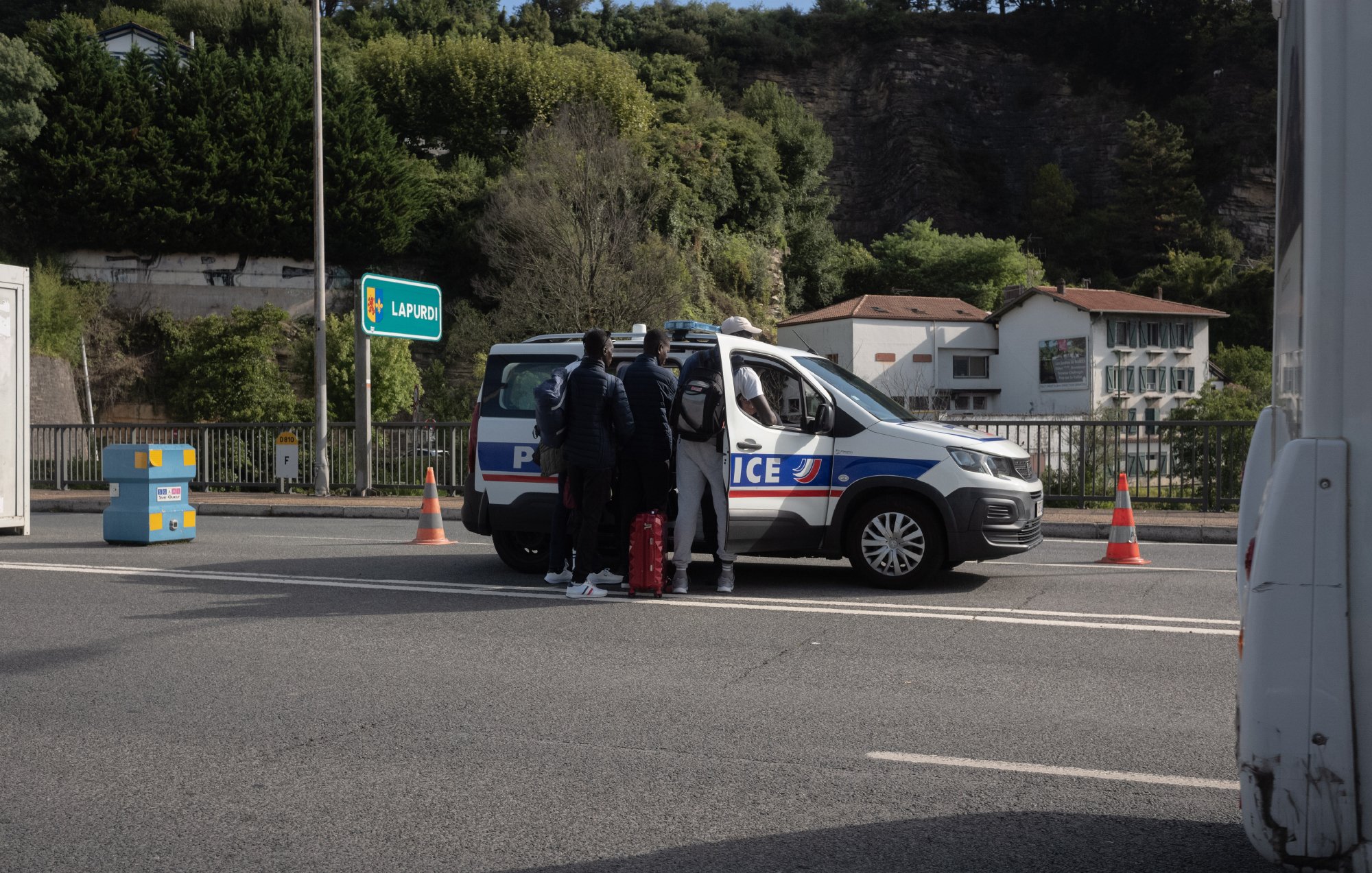 L'image montre une scène sur une route, probablement un contrôle de police. Au centre, on voit une voiture de police de couleur grise avec des marquages bleus et rouges, arrêtée sur la route. Quatre personnes se tiennent près de la voiture, dont deux semblent discuter avec les agents. Ils portent des vêtements sombres et certains semblent avoir des bagages. En arrière-plan, on aperçoit des bâtiments blancs et des arbres qui bordent la route, avec un panneau indiquant "LARRESO". Des cônes de signalisation orange sont disposés autour de la voiture pour sécuriser la zone. L'ambiance paraît calme, bien que la situation soit formelle.