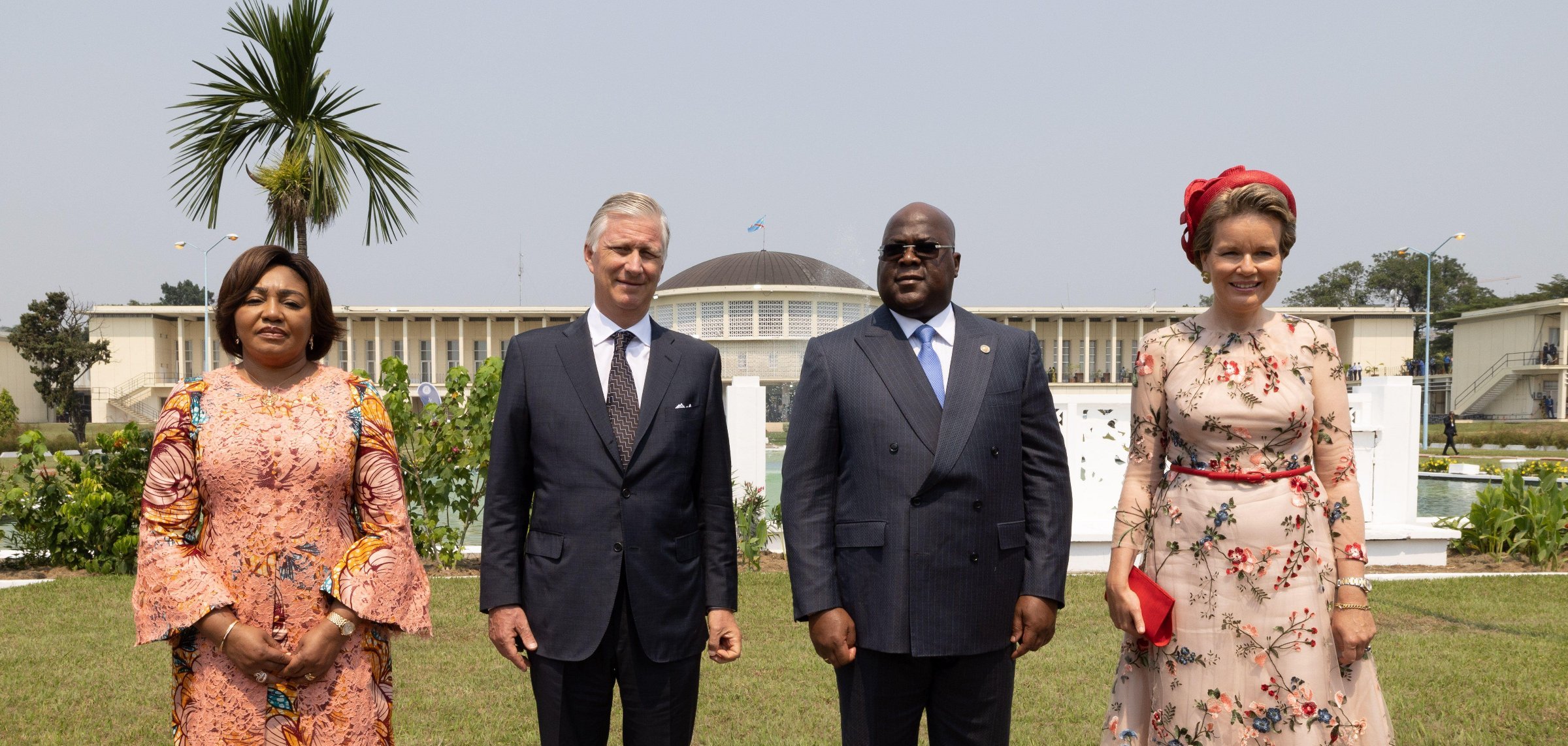 L'image montre un groupe de quatre personnes se tenant debout dans un jardin bien entretenu. À gauche, une femme porte une robe longue à motifs floraux et des manches évasées. À côté d'elle, un homme en costume sombre est debout, affichant une expression chaleureuse. Ensuite, un autre homme, également en costume, a une posture amicale et est au centre de l'image. Finalement, à droite, une femme porte une robe à fleurs avec un joli chapeau assorti et un sac à main rouge, se tenant avec élégance. En arrière-plan, on aperçoit un bâtiment imposant et un palmier qui ajoutent une atmosphère exotique à la scène. Le ciel est clair, ce qui contribue à un sentiment de rencontre officielle.