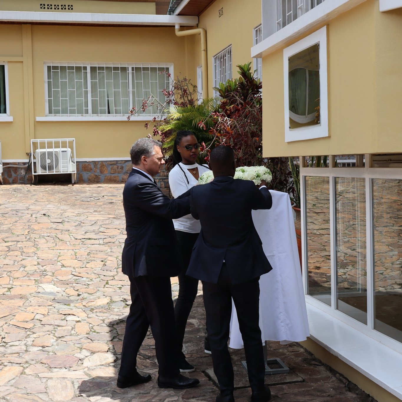 Dans cette image, nous voyons un événement en extérieur, probablement une cérémonie de dévoilement. Trois hommes sont présents. L'un d'eux, portant un costume sombre, est en train de tirer une couverture blanche qui cache un objet ou une plaque. À ses côtés, un homme en tenue décontractée tient un bouquet de fleurs. En arrière-plan, on peut apercevoir un bâtiment avec de grandes fenêtres et une pelouse soignée, créant une atmosphère accueillante. Le sol est en pierre, ajoutant un aspect naturel à l'environnement. L'ensemble de la scène évoque un moment de célébration ou d'inauguration.