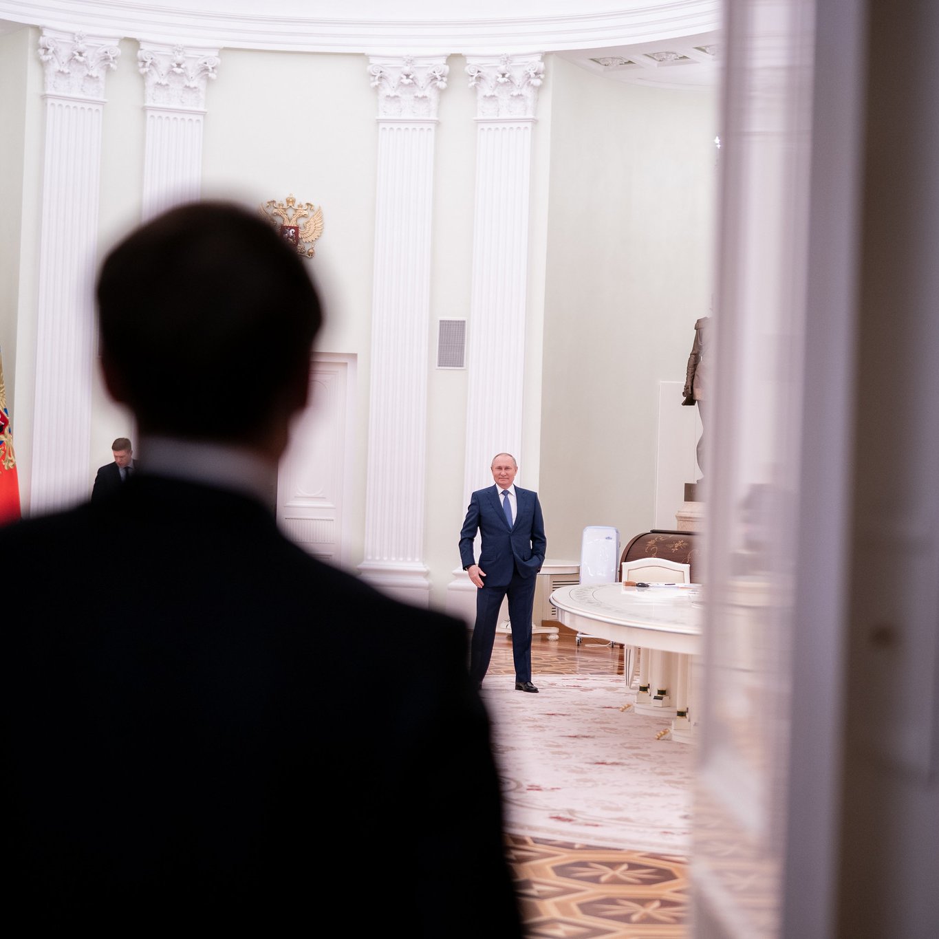 L'image montre une scène dans un intérieur officiel, probablement un palais ou un bureau gouvernemental. Au premier plan, on voit le dos d'une personne en costume sombre, qui semble faire face à une autre personne située à une certaine distance. Cette deuxième personne, qui est clairement identifiable comme une figure politique importante, se tient debout dans une pièce au décor élégant, avec une lumière douce. À l'arrière-plan, on peut distinguer des drapeaux russe et peut-être d'autres éléments symboliques. Le sol est en marbre, et les murs sont peints dans des teintes claires, ajoutant une ambiance solennelle à l'ensemble. L'atmosphère de l'image suggère un moment de rencontre ou de dialogue important.