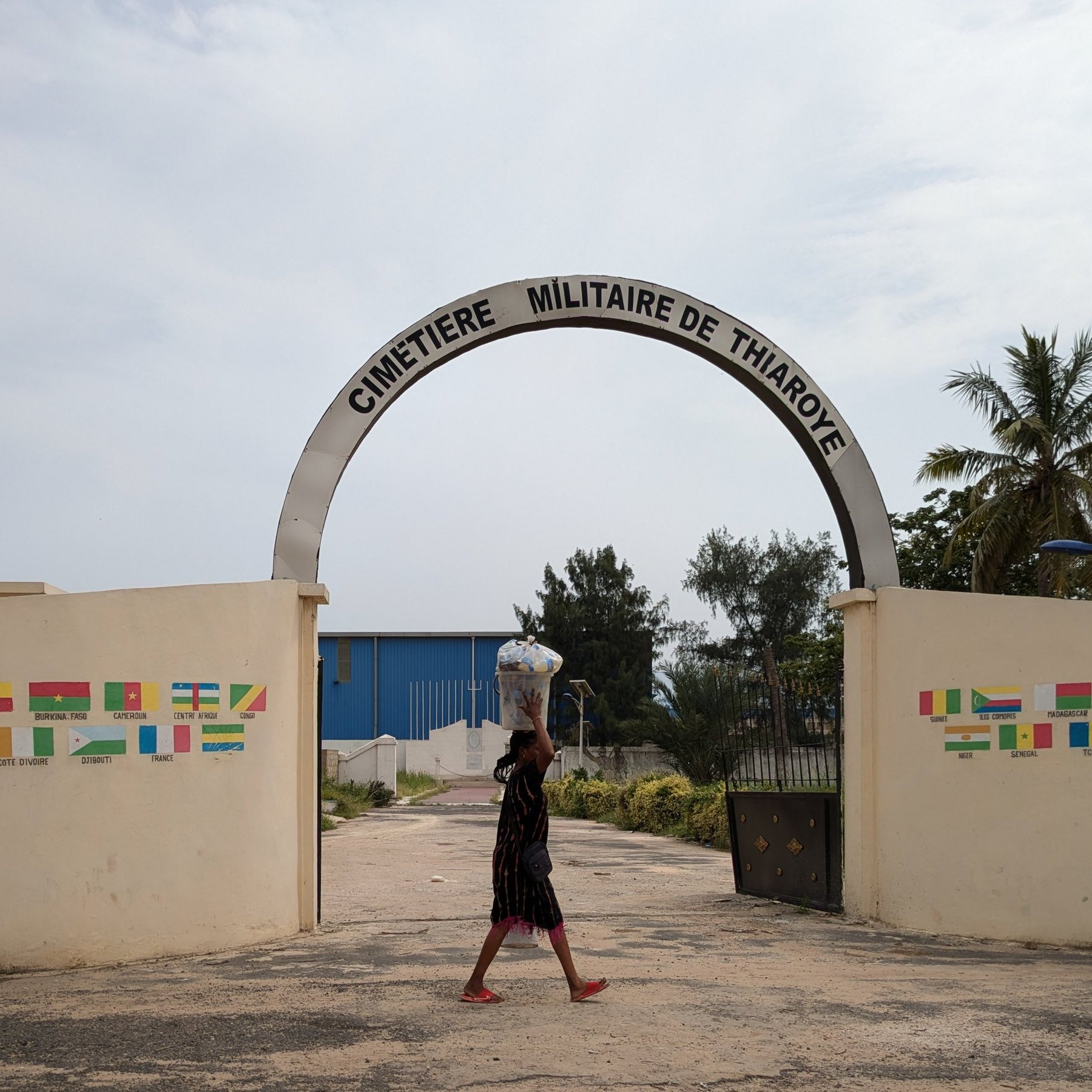 L'image montre l'entrée du cimetière militaire de Thiaroye. Un grand arc de portail est inscrit avec le nom "CIMETIÈRE MILITAIRE DE THIAROYE". En arrière-plan, on peut apercevoir quelques arbres, dont des palmiers, qui ajoutent une touche tropicale à la scène. Au premier plan, une personne marche, portant un grand récipient sur sa tête. Le sol est en béton, avec des traces de circulation. De part et d'autre de l'entrée, des murs affichent des drapeaux de différentes nations, apportant une sensation de diversité. L'ensemble dégage une ambiance calme et respectueuse.
