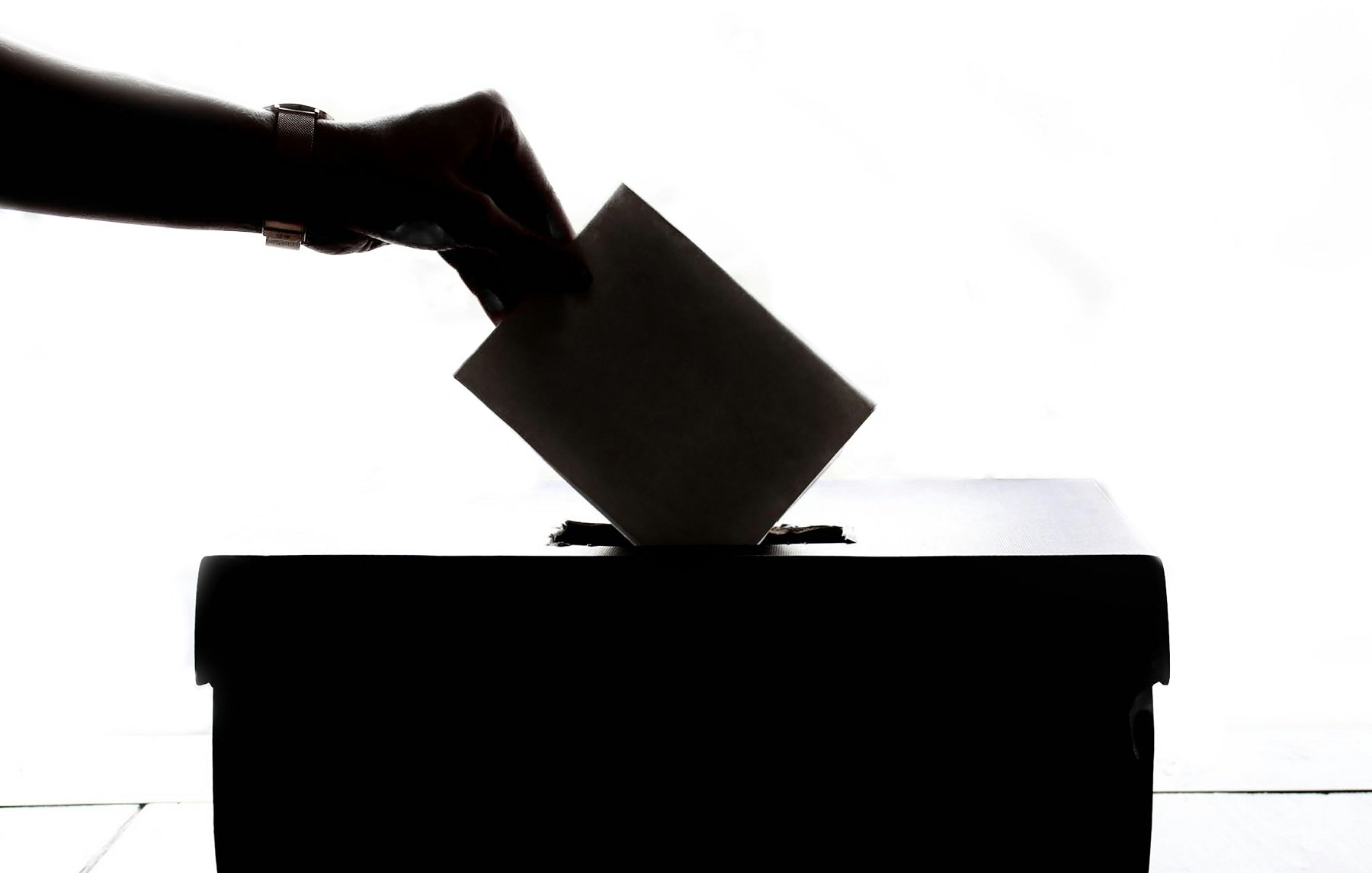 L'image représente une scène de vote. On peut voir une main tendue vers une urne, prête à y insérer un bulletin de vote. La silhouette de la main et de l'urne est visible contre un fond lumineux, créant un contraste fort. L'urne est de forme rectangulaire et se trouve sur une surface plane. Cette image évoque le processus démocratique et l'importance de l'engagement civique.