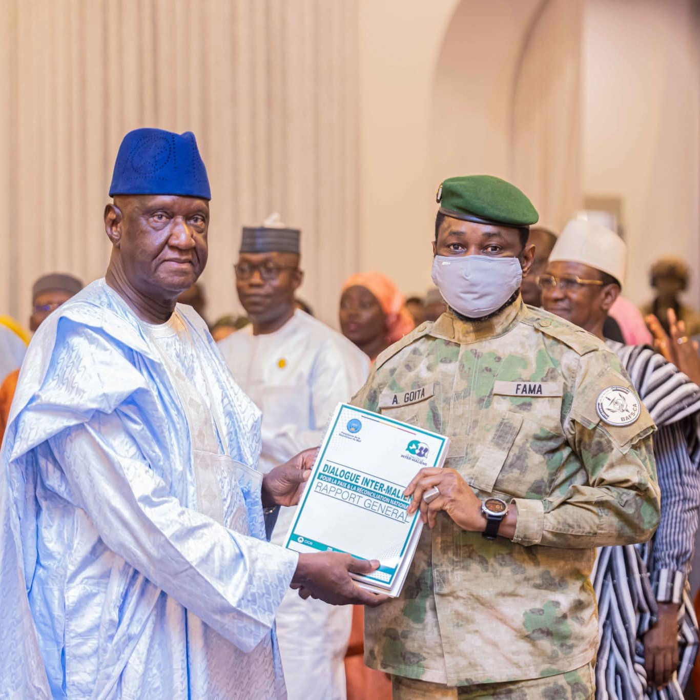 L'image montre un événement officiel ou une cérémonie de remise de prix. Au premier plan, un homme portant une tenue traditionnelle, composée d'une longue robe et d'un chapeau bleu, reçoit un document d'un officier militaire. Ce dernier est vêtu d'un uniforme camouflé et porte un masque gris. En arrière-plan, plusieurs personnes en tenue variée assistent à l'événement, certaines applaudissant, tandis que d'autres observent. L'environnement semble formel, avec des décorations adaptées à l'occasion.