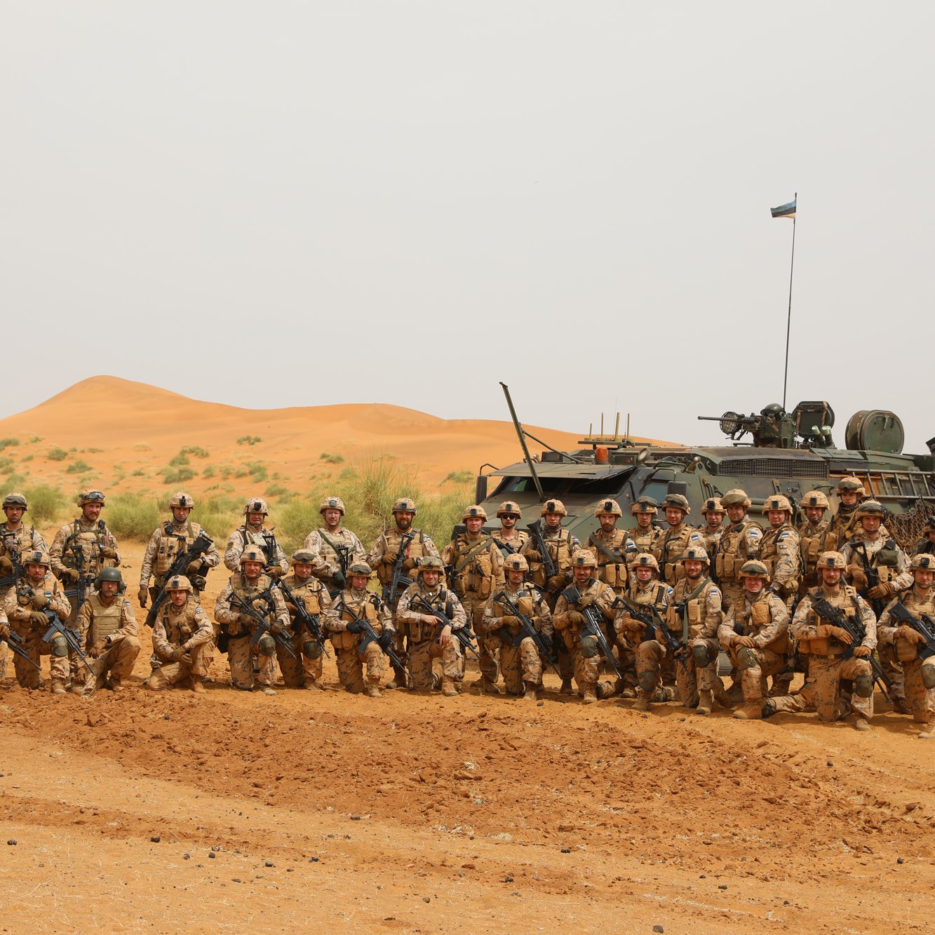 L'image montre un groupe de soldats rassemblés dans un paysage désertique. Au premier plan, on peut voir une trentaine de militaires, tous vêtus de tenues camouflées dans des tons de sable et de marron. Ils se tiennent debout ou accroupis, posant avec des expressions sérieuses. À l'arrière, un véhicule militaire, probablement un blindé, est stationné et porte un drapeau sur son toit. Le fond est marqué par des dunes de sable orangé, typiques des déserts, sous un ciel clair. Cette scène évoque un contexte militaire et une atmosphère de camaraderie entre les soldats.