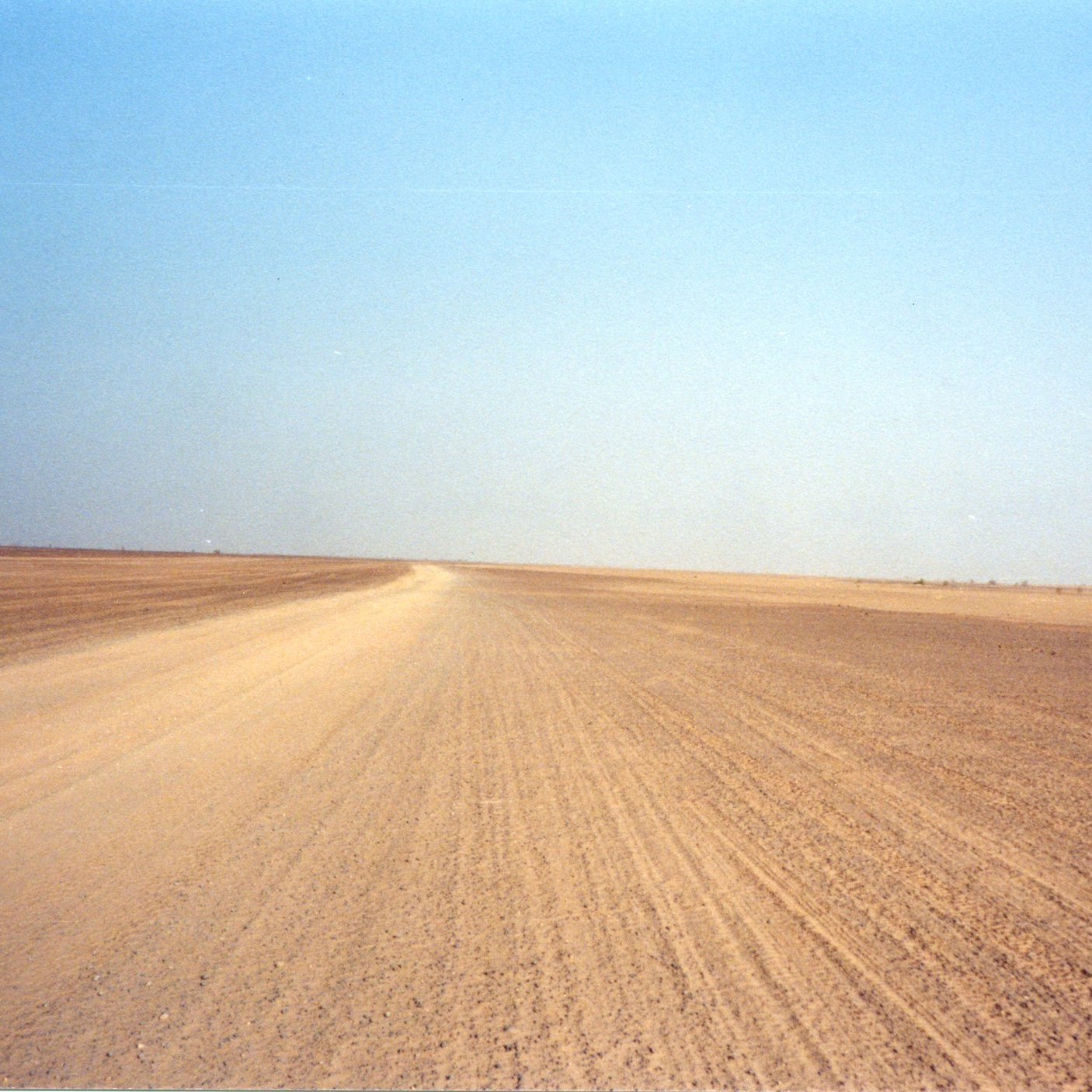 The image depicts a barren landscape characterized by a vast, empty stretch of land, likely a desert or arid region. The foreground features a dirt road that leads into the distance, flanked by dry, flat terrain. The sky overhead is clear and blue, creating a stark contrast with the earthy tones of the ground. The overall scene conveys a sense of isolation and openness, typical of remote, dry environments.
