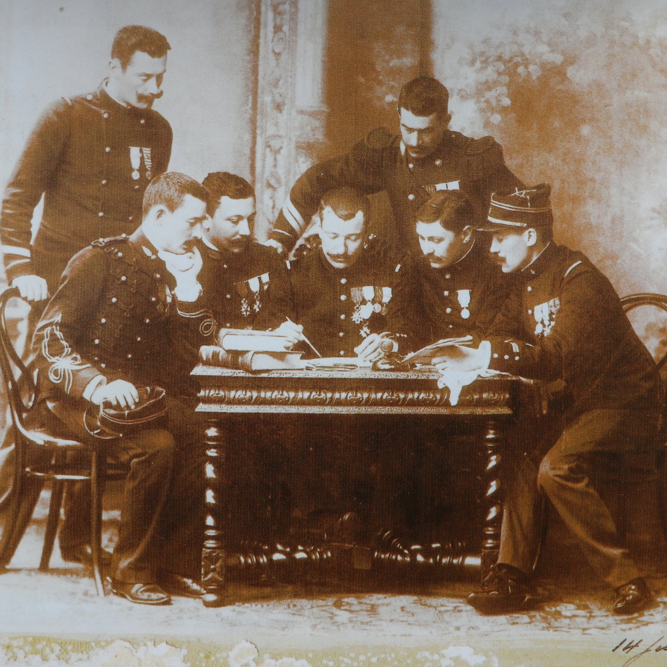Cette image en noir et blanc représente un groupe de six hommes en uniforme, assis autour d'une table en bois. Ils semblent être des membres d'un corps militaire, chacun portant des médailles décoratives sur leur uniforme. Les hommes sont concentrés sur un document ou un plan posé sur la table, et leur posture indique un échange d'idées ou de discussions sérieuses. En arrière-plan, on peut distinguer quelques éléments architecturaux flous, suggérant un intérieur de bâtiment classique. L'atmosphère se dégage une certaine solennité et camaraderie.