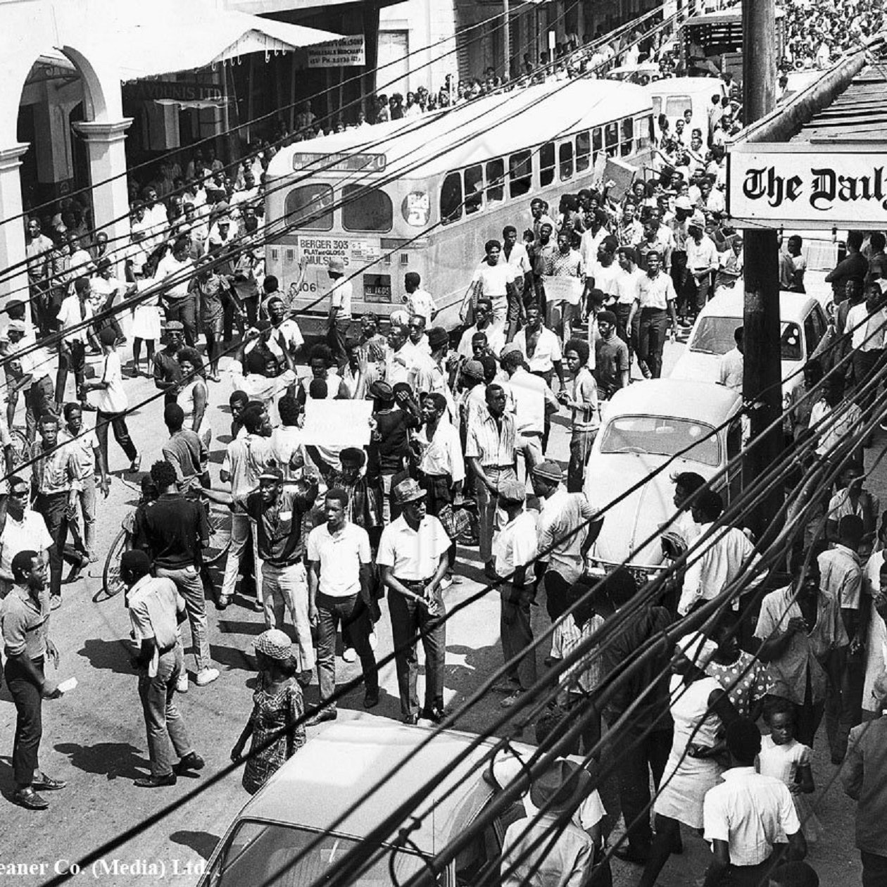 L'image que vous avez devant vous représente une scène urbaine animée dans les années 1960. On peut voir une foule dense de personnes marchant dans une rue. Les gens portent des vêtements typiques de l'époque, et il semble qu'ils se déplacent avec un but précis. À gauche, plusieurs commerces sont visibles, avec des enseignes en lettres visibles. Un ancien bus à l'allure caractéristique est garé, ajoutant une touche nostalgique à la scène. On aperçoit également quelques voitures stationnées et d'autres qui circulent dans la rue. Les fils électriques sont visibles en haut de l'image, accentuant l'impression d'une ville vivante et dynamique. L'atmosphère générale évoque une période de mouvement socioculturel important, avec des échanges et une vitalité palpable dans l'air.