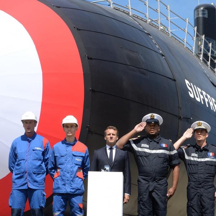 L'image représente un sous-marin de type Suffren, mis en avant lors d'une cérémonie en extérieur. Le sous-marin, aux couleurs caractéristiques bleu, blanc et rouge, se dresse majestueusement avec une grande coque stylisée. Le chiffre « 12 » est visible sur le côté. Devant le sous-marin, on voit plusieurs personnes en tenue de travail. À gauche, deux hommes portent des combinaisons bleues, un troisième homme, en costume, semble féliciter le projet, tandis que le quatrième, en uniforme, adopte une posture militaire. L'arrière-plan est dégagé avec un ciel bleu, ce qui donne une impression de journée ensoleillée. La scène évoque un moment de fierté et de célébration pour les employés et les responsables militaires, symbolisant un jalon important dans le domaine naval.